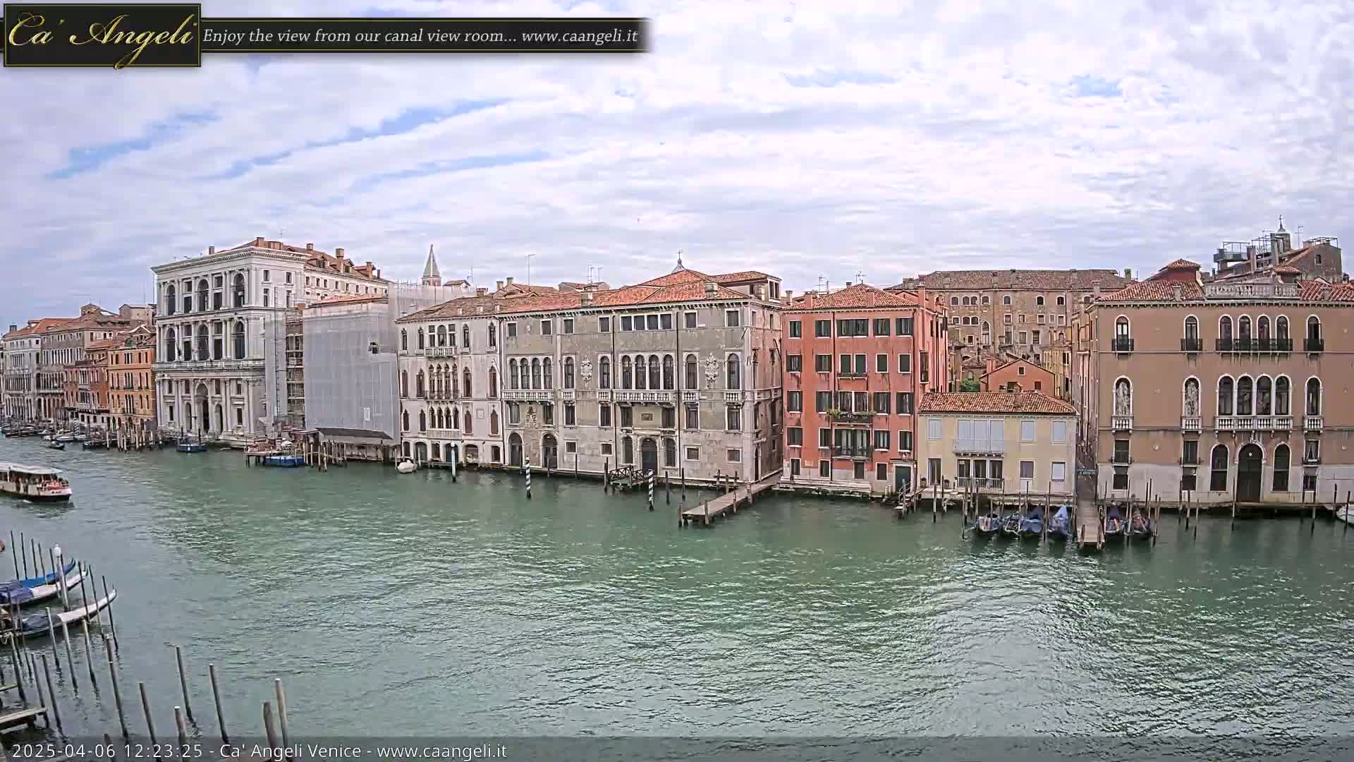 A canal in Venice, under a partly cloudy sky, is lined with several historic buildings, gondolas, and boats.