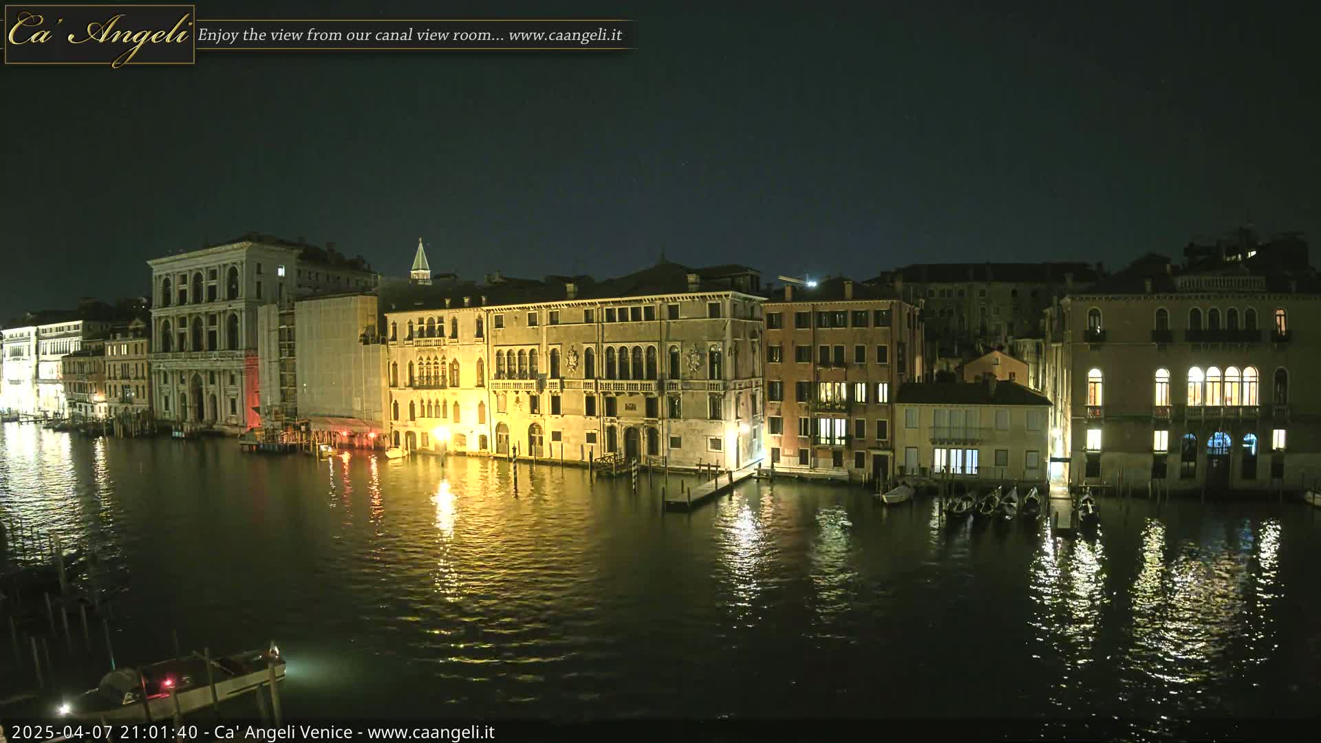 A nighttime view of a canal in Venice, Italy, showing illuminated buildings along the water's edge and several boats moored.