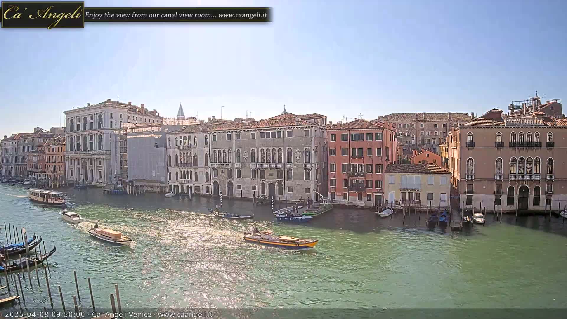 Under a bright, sunny sky, several boats travel along a canal lined with historic Venetian buildings.