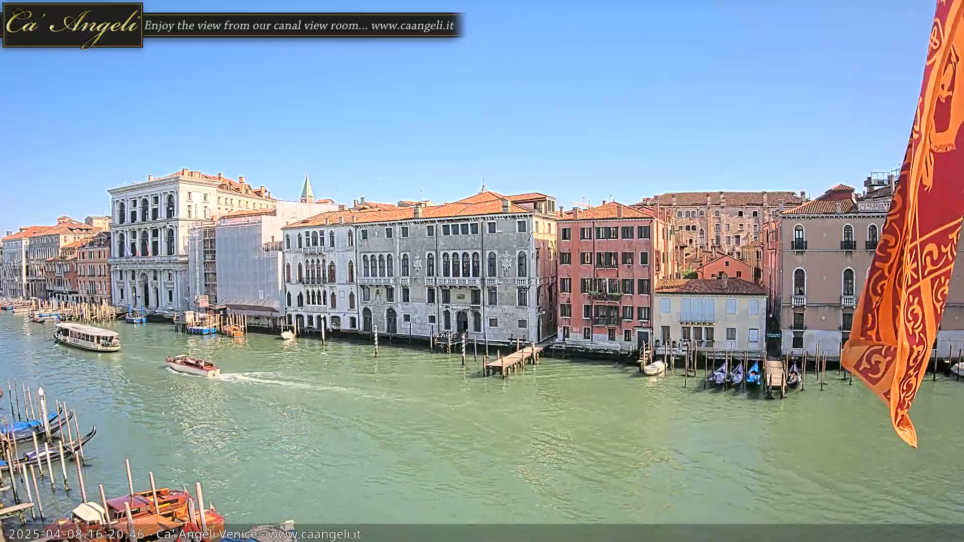 A sunny day in Venice shows a canal with boats, gondolas, and several multi-story buildings lining the waterway.