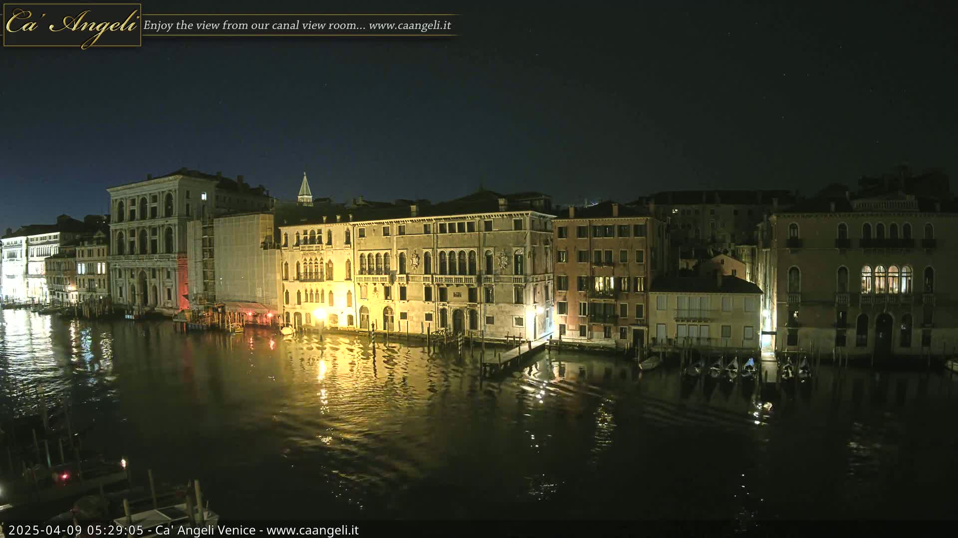 A nighttime view of a canal in Venice, Italy, shows several buildings lit up along the waterway, with gondolas moored along the banks.