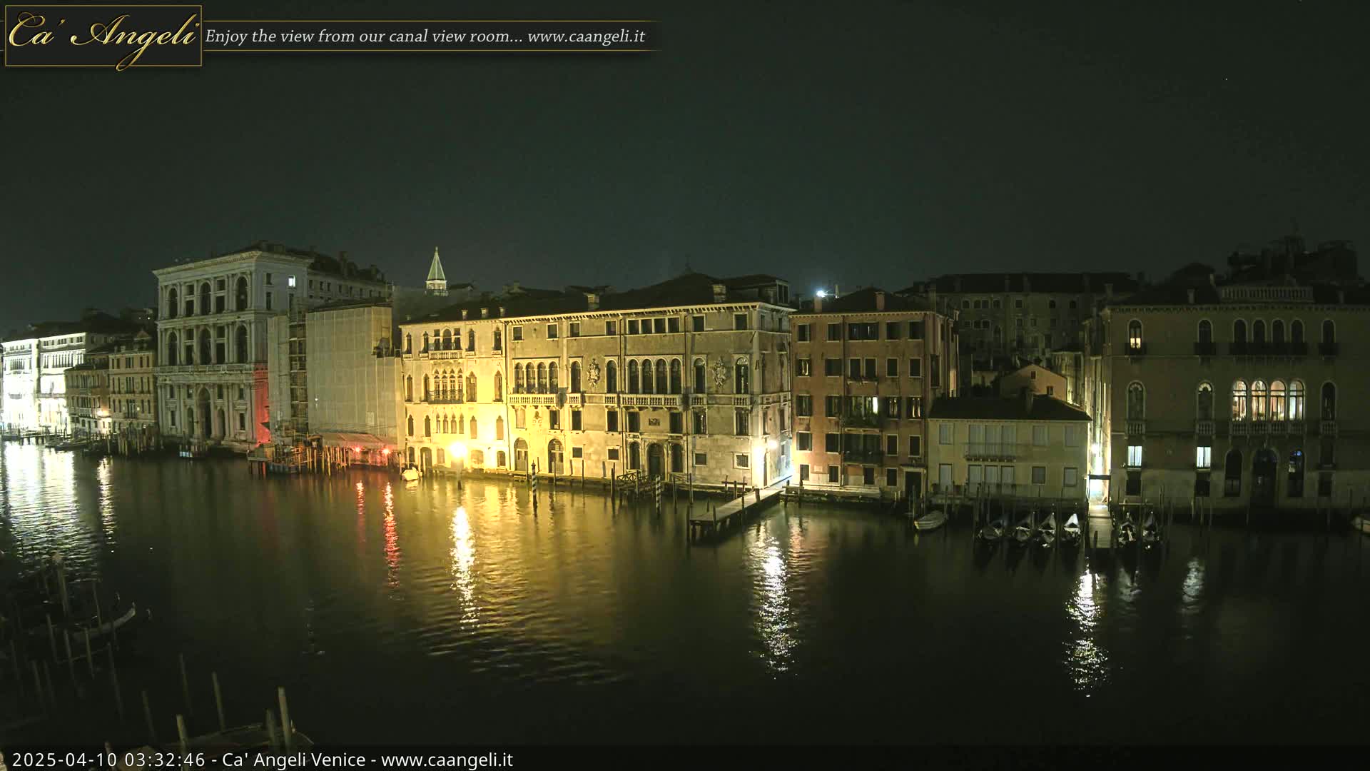 A nighttime view of illuminated buildings lining a canal, with several gondolas moored along the water's edge.