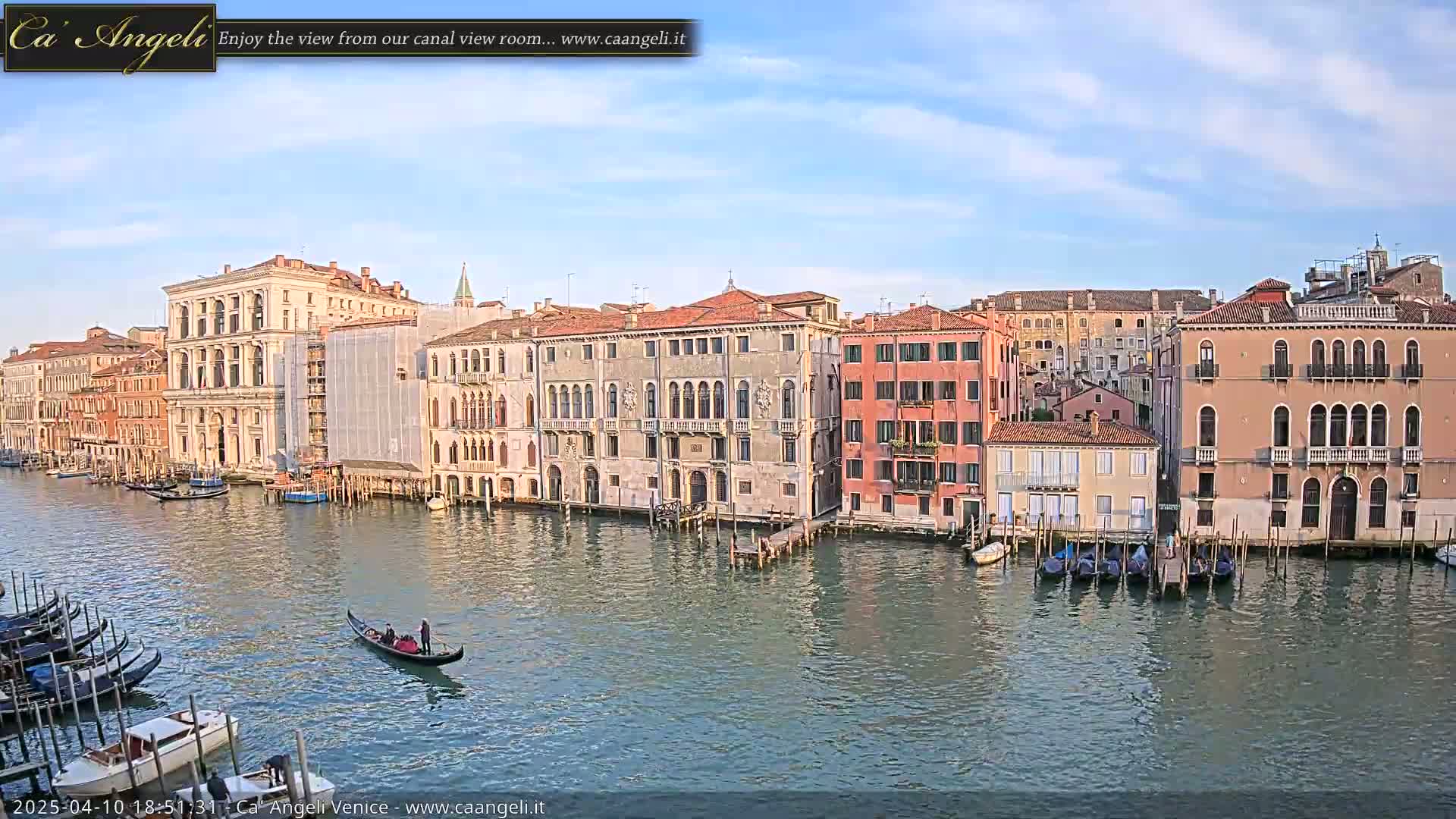 A gondola glides along a calm canal in Venice, under a partly cloudy sky, past several historic buildings lining the waterway.
