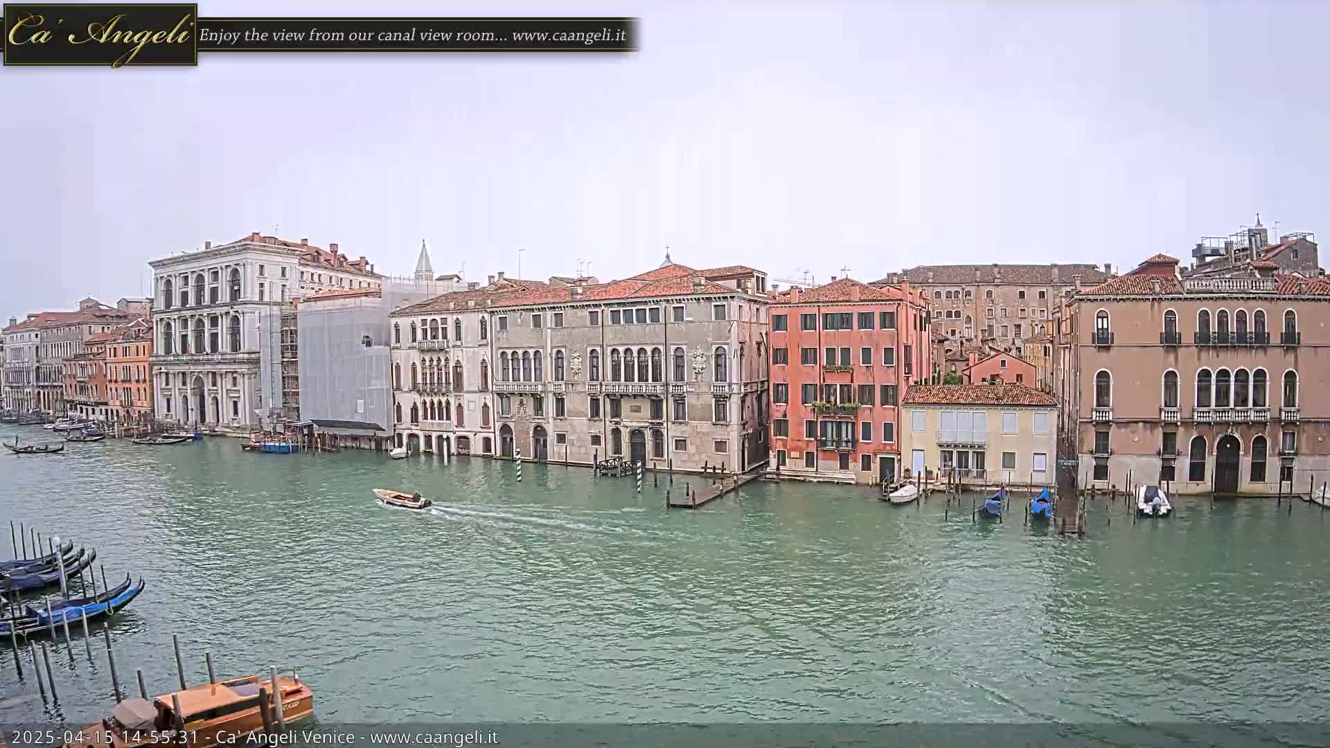 A canal in Venice, Italy, with several gondolas and motorboats, is viewed from across the water on an overcast day; many historic buildings line the canal.