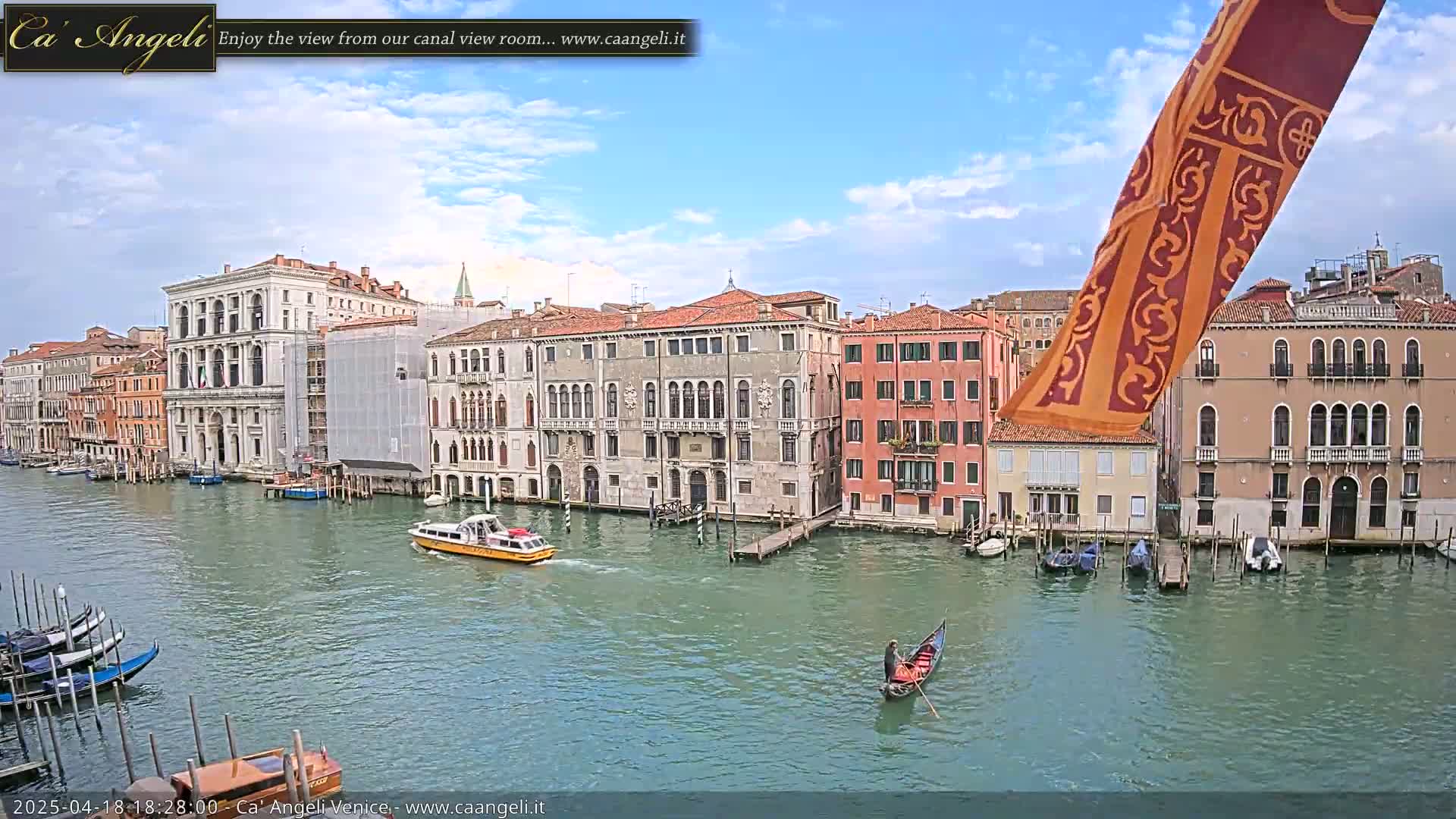 A gondola and a motorboat travel on a canal lined with historic Venetian buildings under a partly cloudy sky.