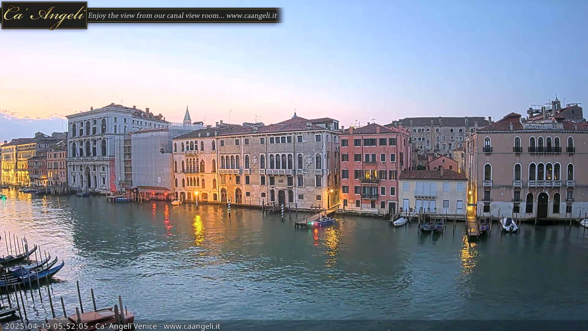 A tranquil canal in Venice at dawn, lined with historic buildings reflecting in the calm water, with a few boats moored along the edge.