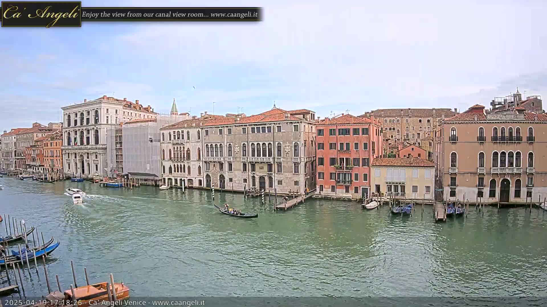 A gondola and several other boats are on a canal in Venice, Italy, under a mostly cloudy sky, with numerous buildings lining the waterway.
