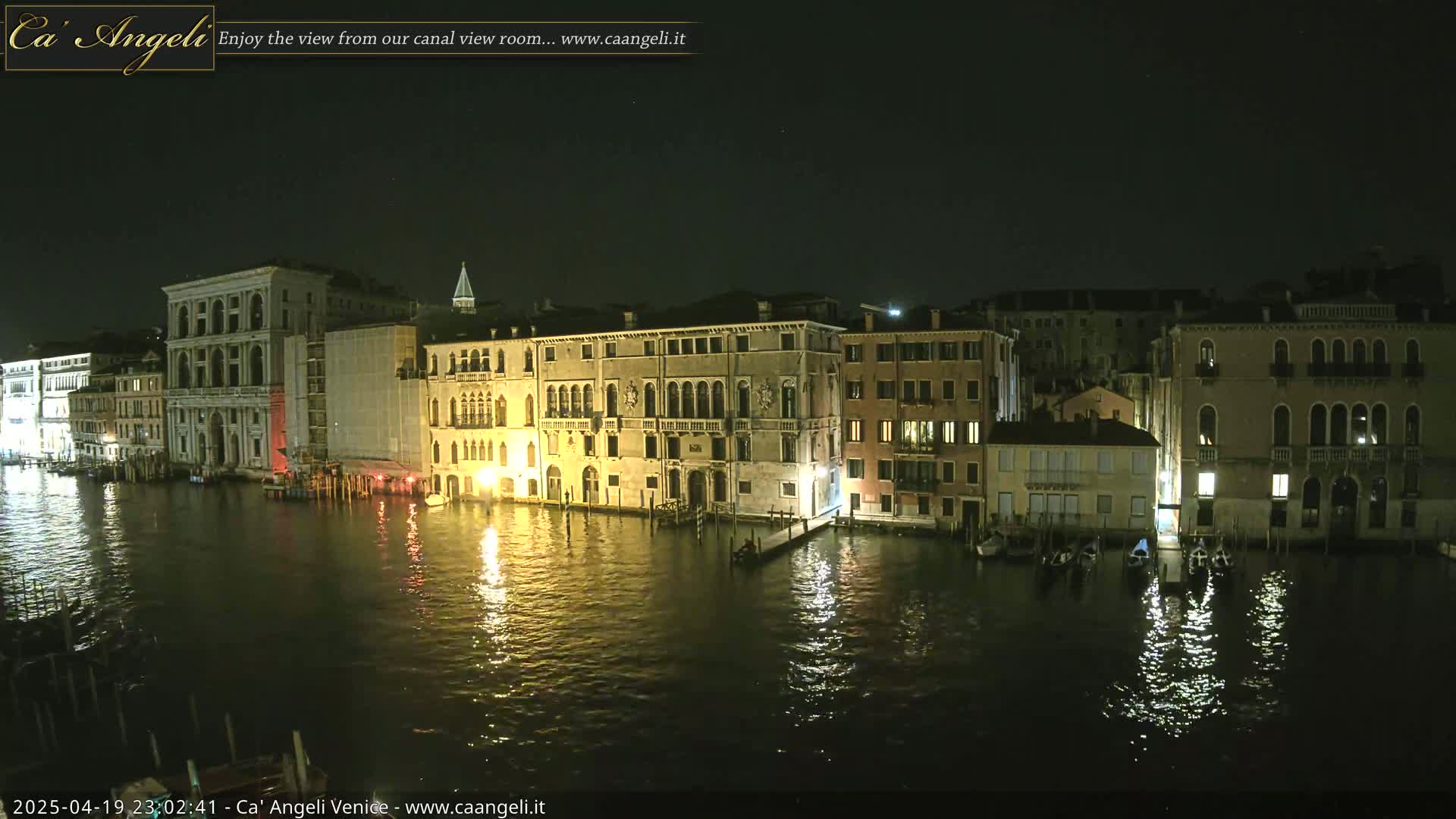 At night, illuminated buildings line a canal, their lights reflecting on the dark water where several gondolas are moored.