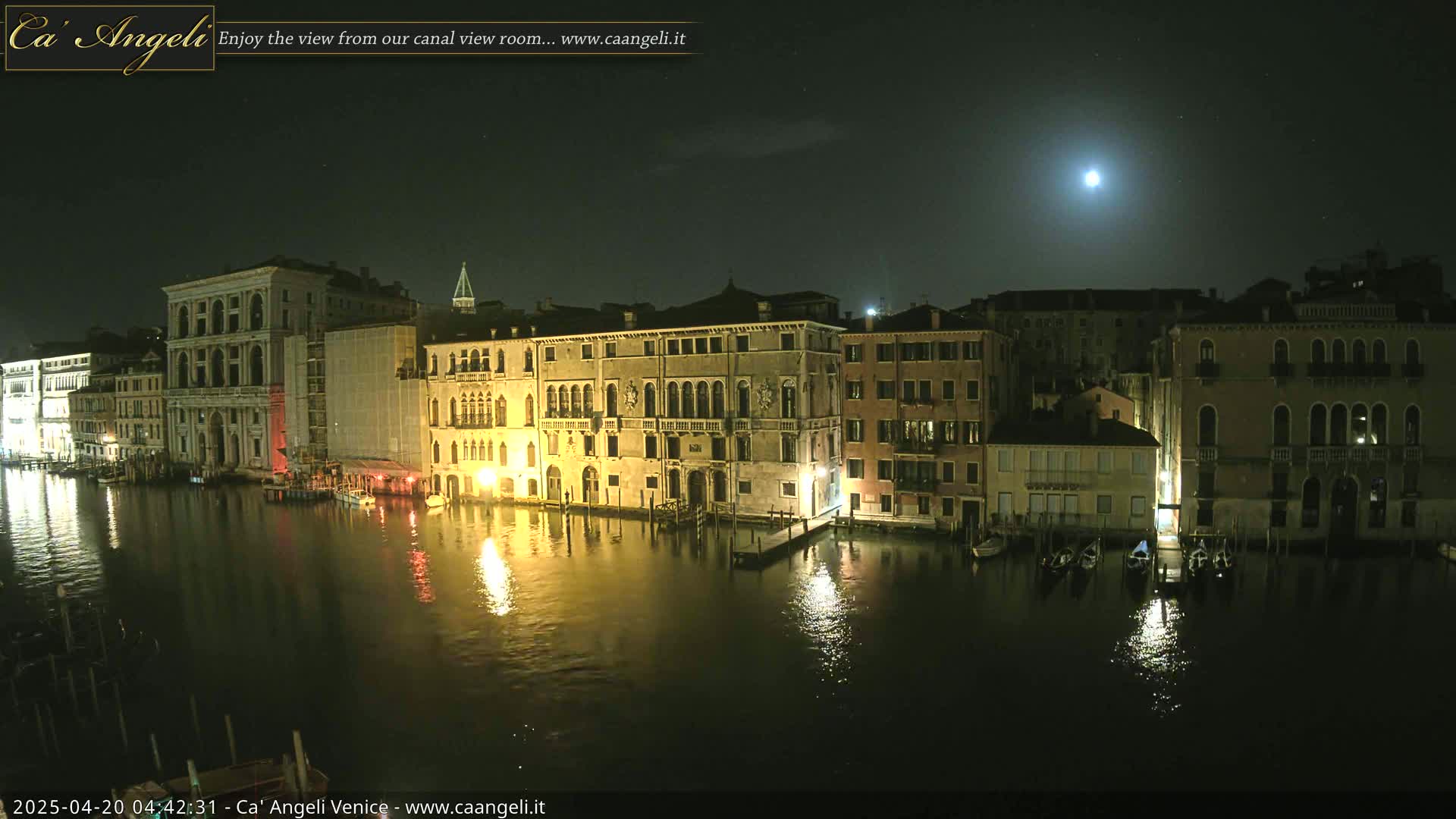 A moonlit night scene shows a canal in Venice with several buildings illuminated along its banks, gondolas moored, and calm reflective water.