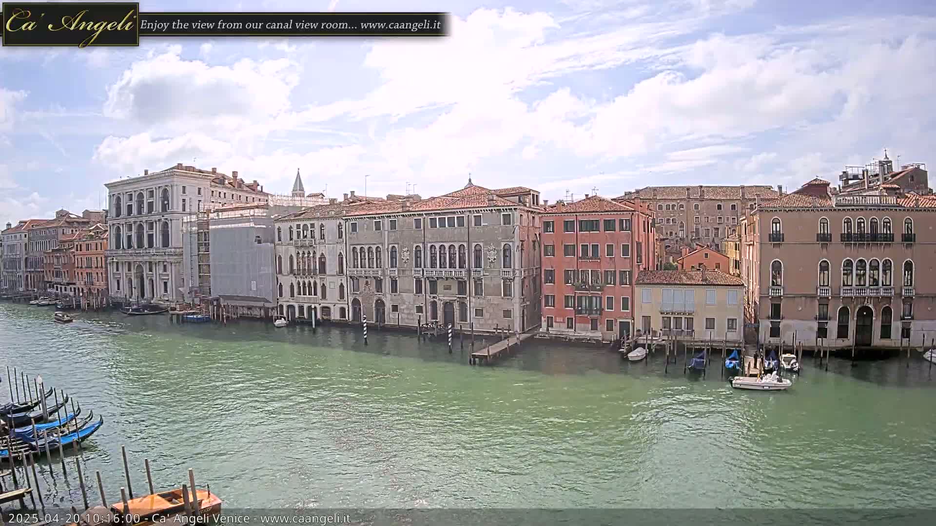 A canal in Venice, under a partly cloudy sky, shows several historic buildings lining its banks, with gondolas and other small boats moored along the water's edge.