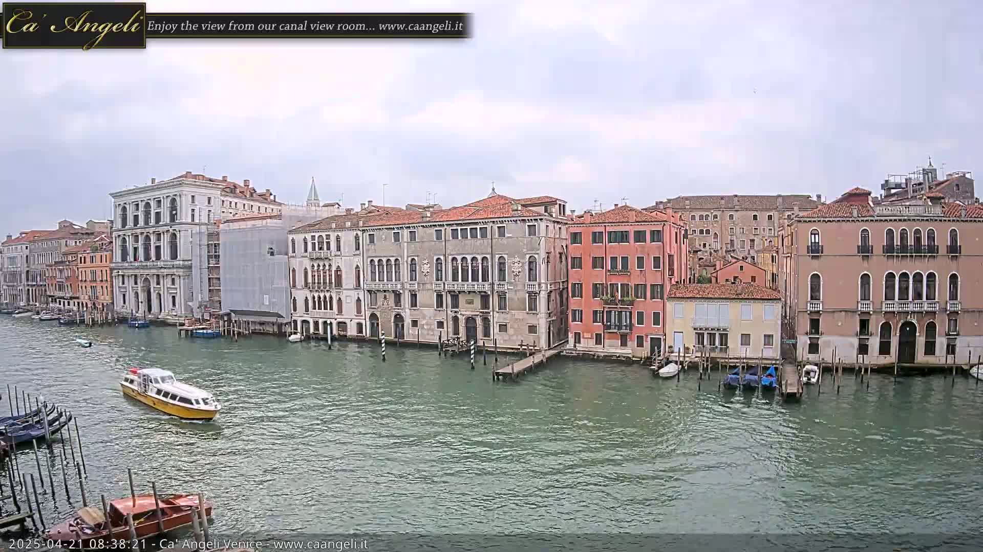 A waterway with several buildings along its edge is shown on an overcast day, with a yellow water taxi traveling across the water.