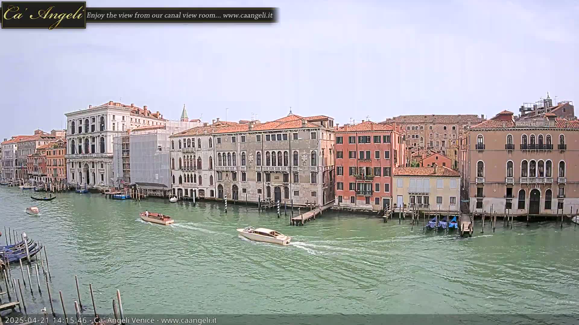 A wide shot shows a calm canal in Venice, Italy, with several boats and numerous buildings lining its banks under a mostly clear sky.