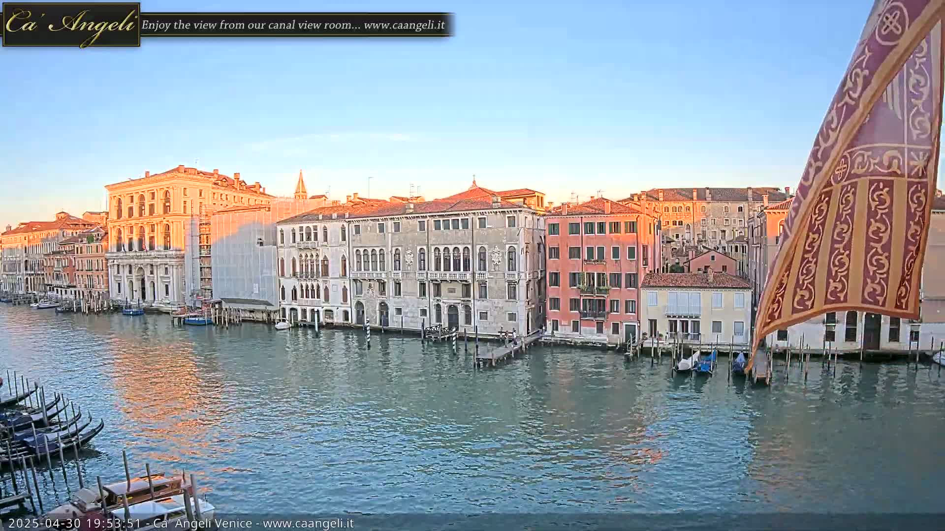 A calm canal in Venice, at sunset, is lined with historic buildings, gondolas are moored along the waterfront, and a patterned flag hangs from a building.