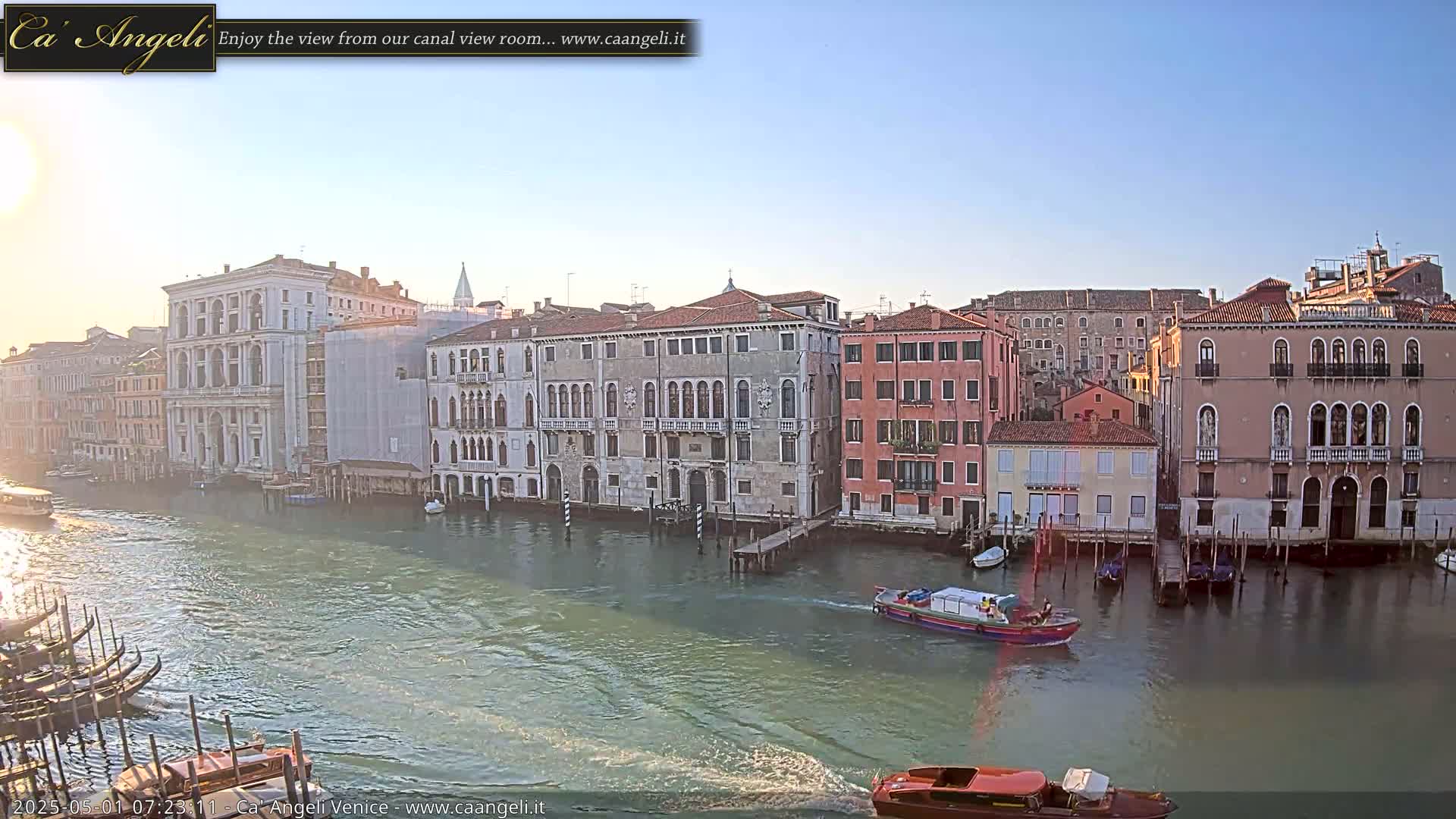 A canal in Venice, under a clear, bright sky, is lined with numerous buildings, with gondolas and motorboats on the water.