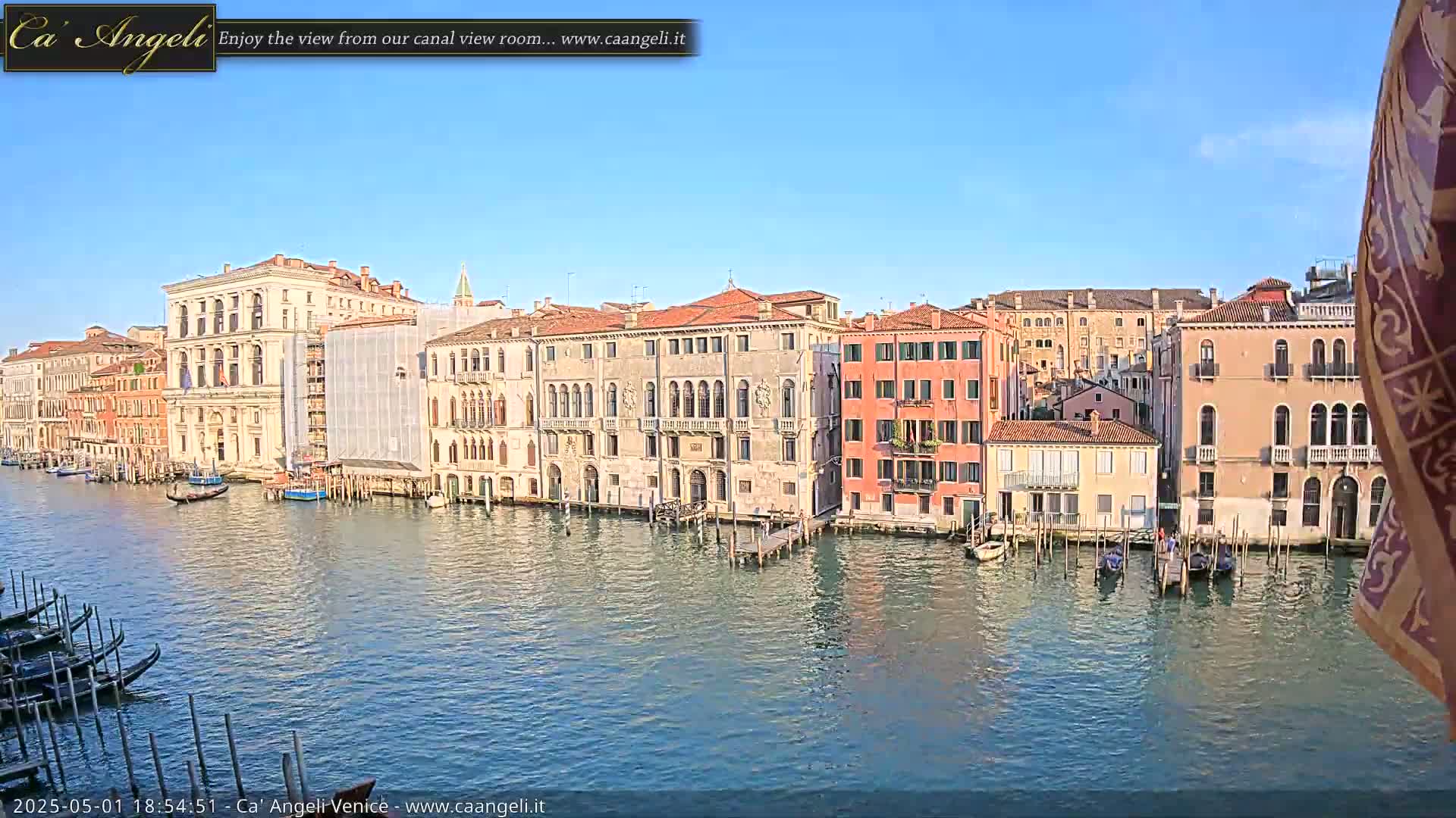 A canal in Venice, under a clear blue sky, is lined with historic buildings and several gondolas are moored along the water's edge.
