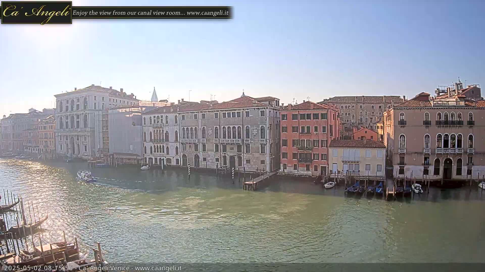 A canal in Venice, Italy, is shown on a sunny day with several gondolas moored along the waterway and several buildings lining the canal.