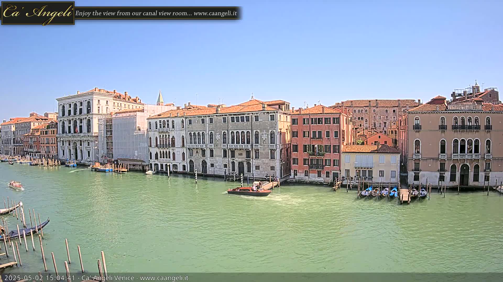 A canal in Venice, under a clear blue sky, is lined with historic buildings, gondolas, and a motorboat.