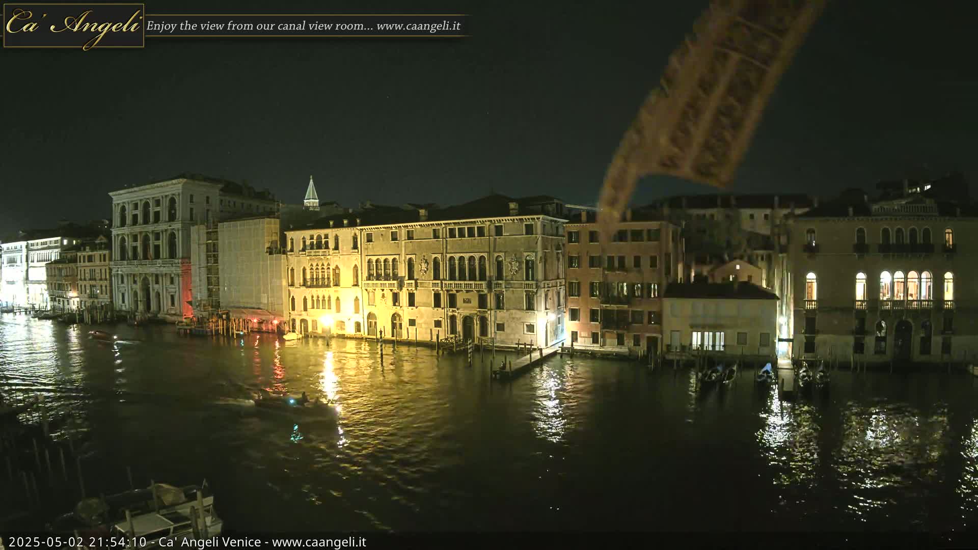 A nighttime view of a canal in Venice, showing illuminated buildings and gondolas on the water under a dark sky.