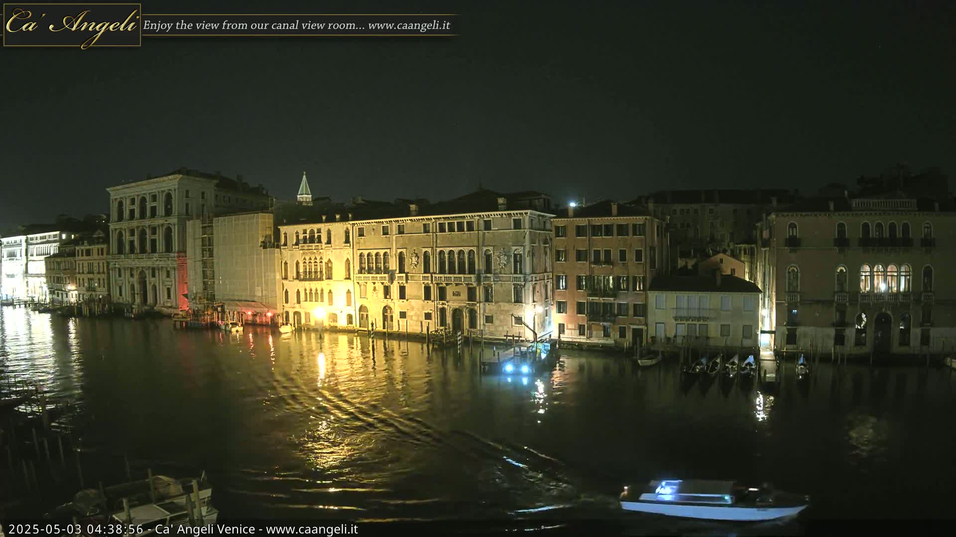 A nighttime view of illuminated buildings lining a calm canal, with a few boats on the water.