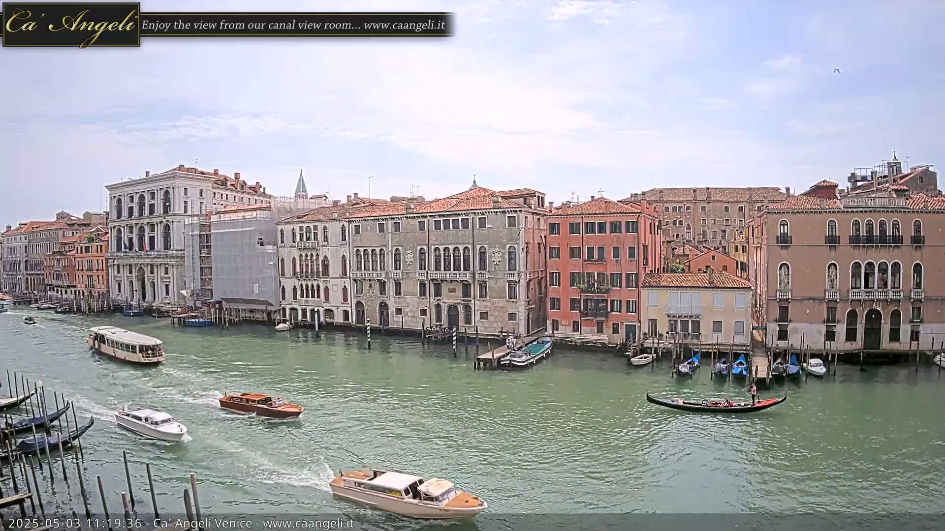A canal in Venice, under a partly cloudy sky, is filled with gondolas and water taxis passing by buildings along the waterfront.