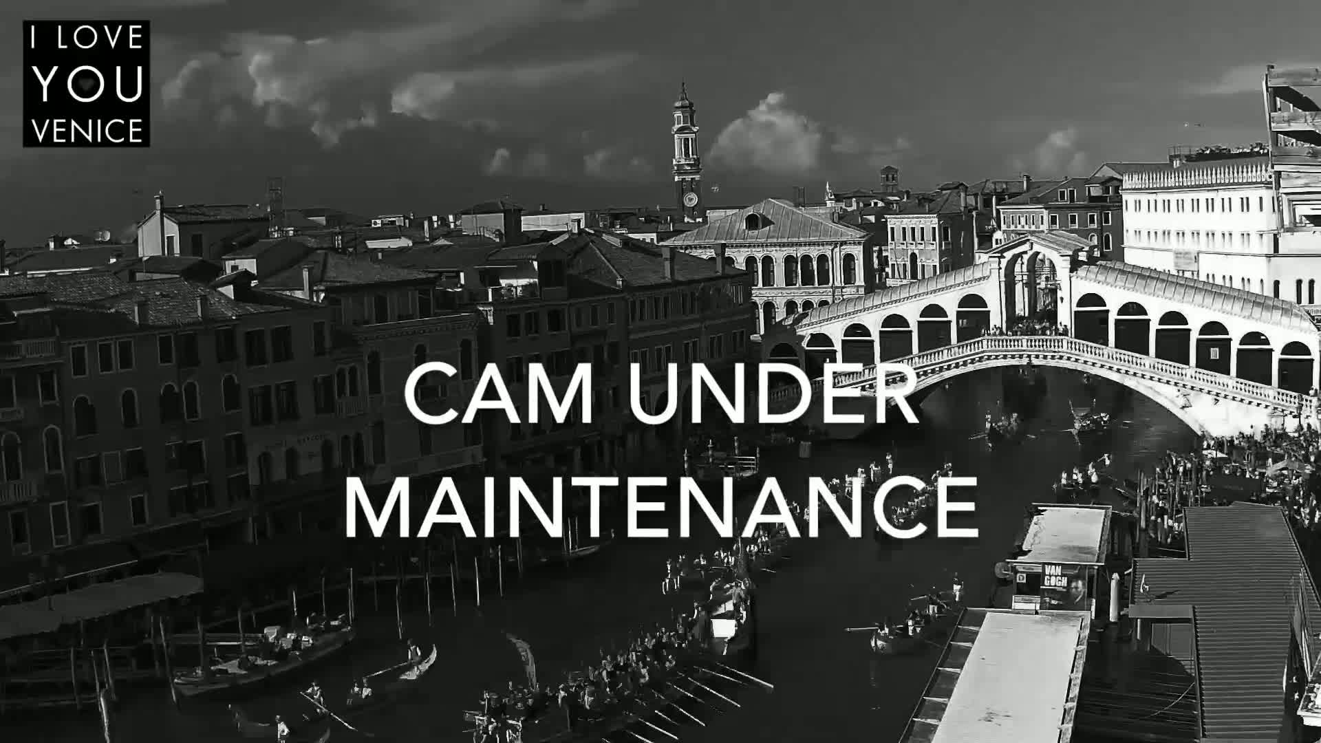 A black and white, high-angle view shows a canal in Venice, Italy, under a partly cloudy sky, with gondolas and other boats on the water, and the Rialto Bridge in the background.