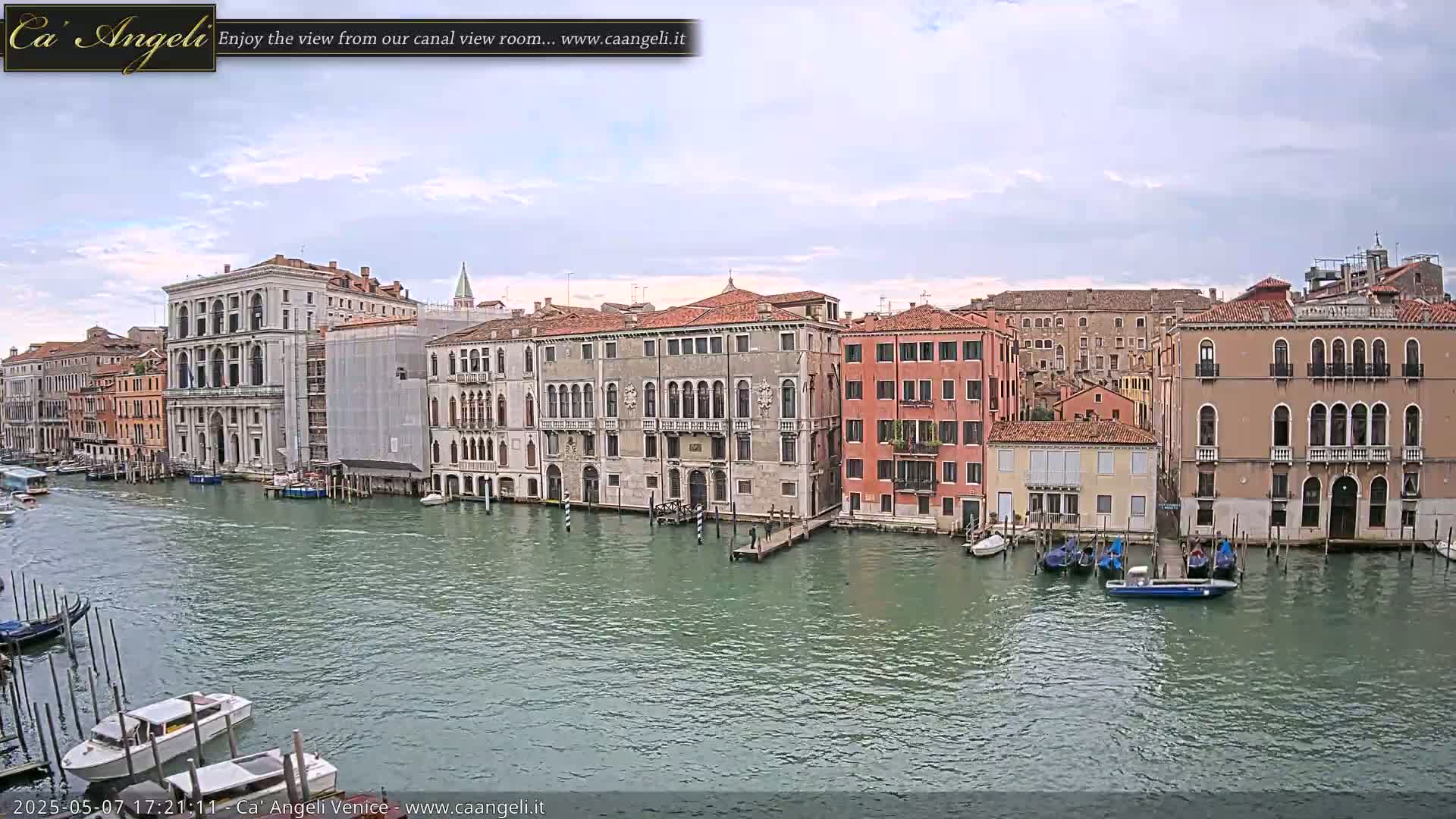 A wide shot of a canal in Venice, Italy, under a partly cloudy sky, shows several large buildings lining the waterway, with numerous boats moored along the shore.