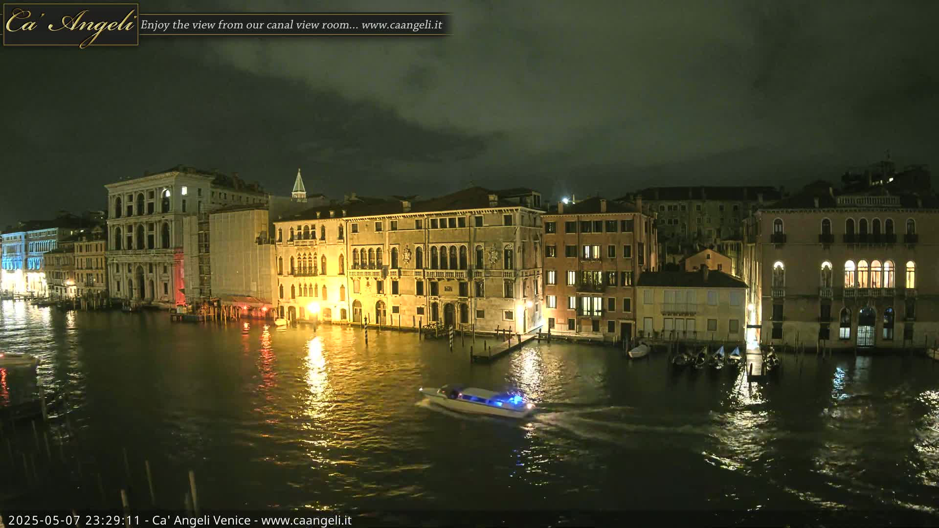 A nighttime view of a canal in Venice, under a dark, cloudy sky, shows illuminated buildings along the water's edge with a small boat passing by.