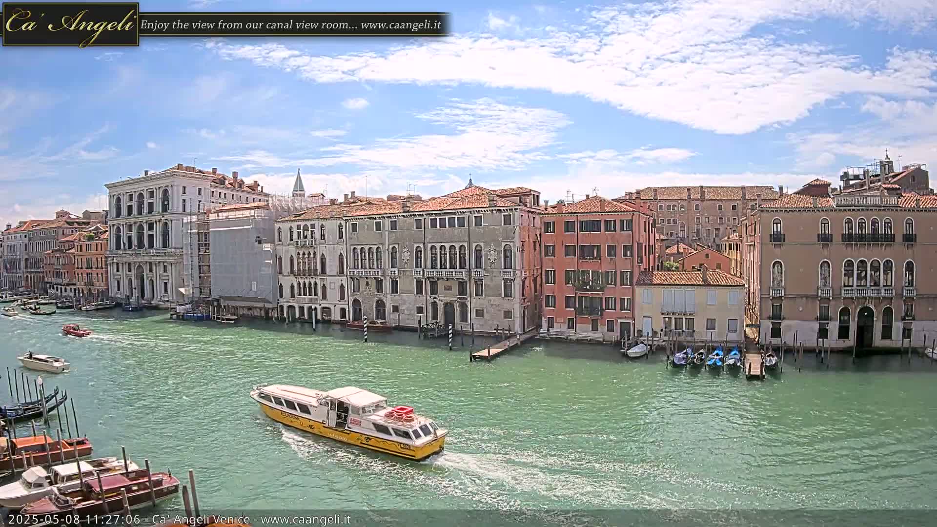 A waterway with several boats, including a yellow water taxi, flows past a row of buildings under a partly sunny sky.