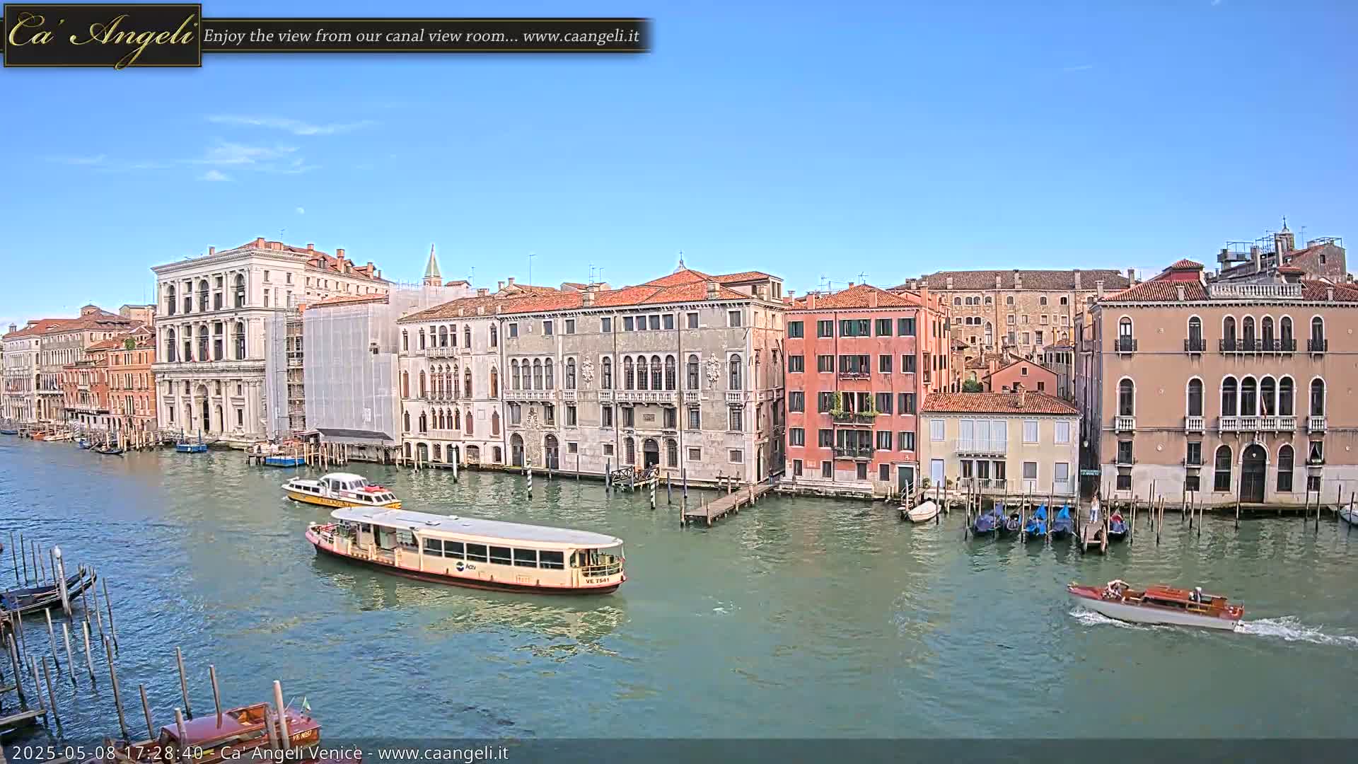A waterway lined with Venetian buildings under a clear blue sky is seen from an elevated viewpoint, with several water vehicles including a passenger ferry and a smaller motorboat navigating the canal.