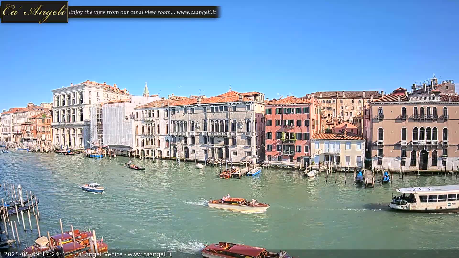 Under a clear blue sky, a wide canal in Venice is filled with boats, and lined with tall, colorful buildings.