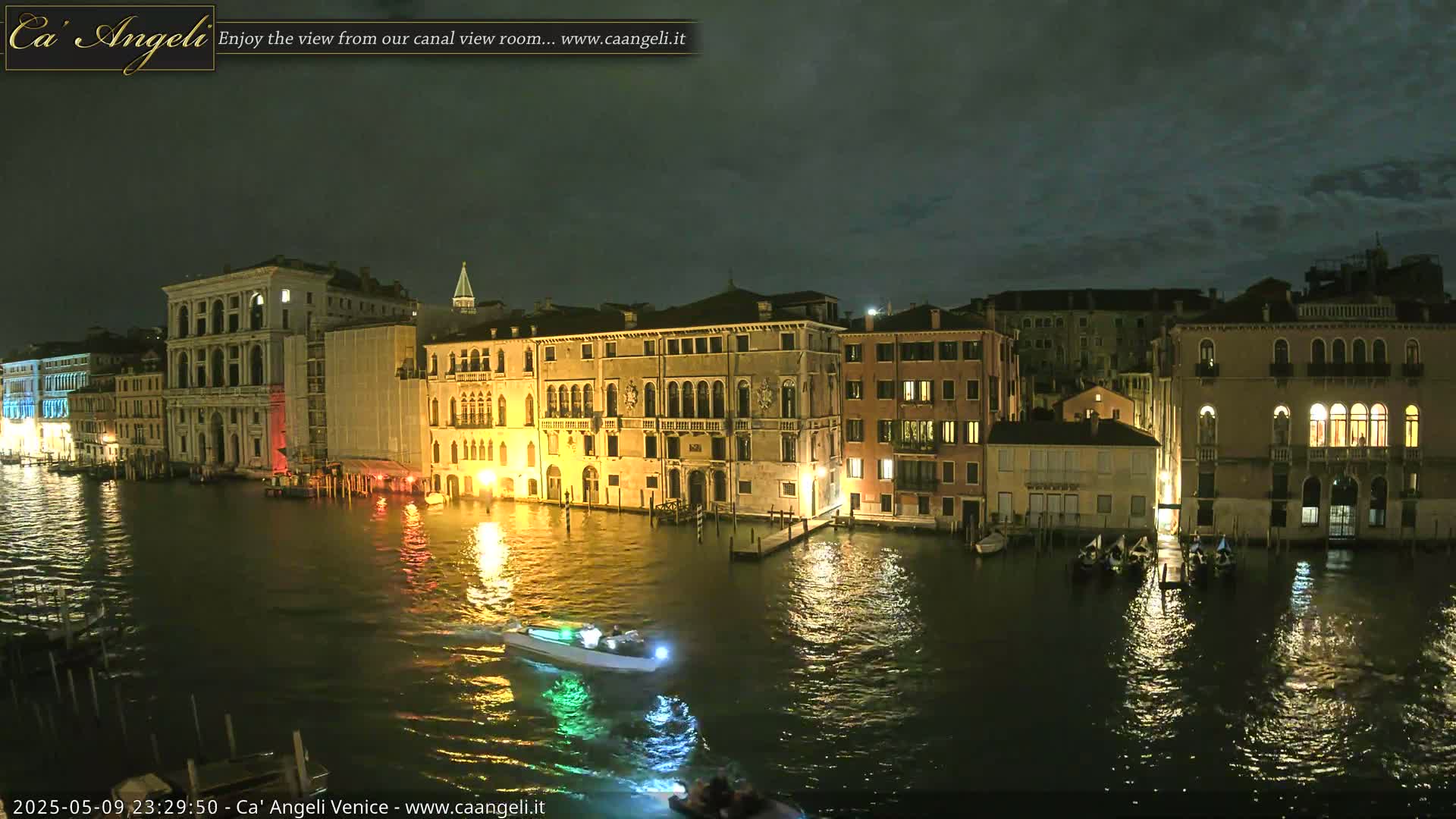 A nighttime view of a canal in Venice, Italy, showing illuminated buildings reflected in dark water, with a small motorboat passing by.