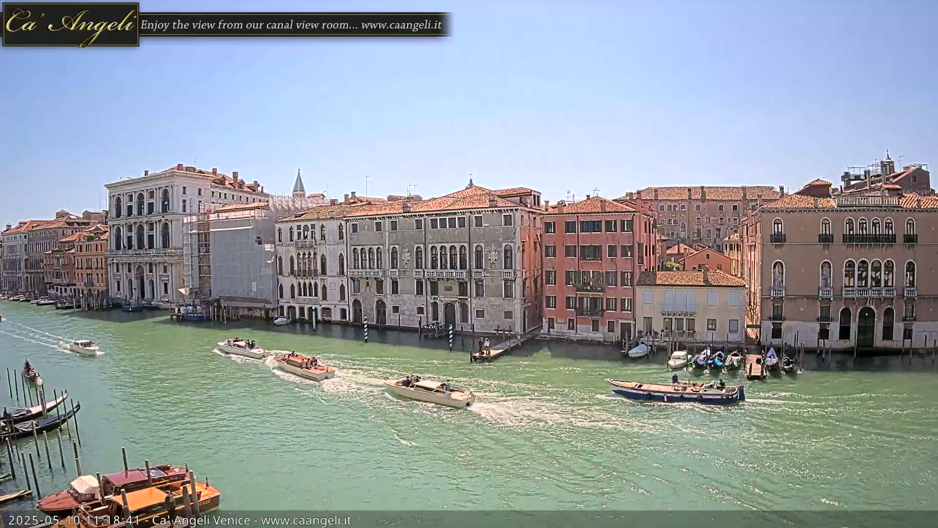 Several motorboats travel on a canal lined with historic buildings under a clear blue sky.