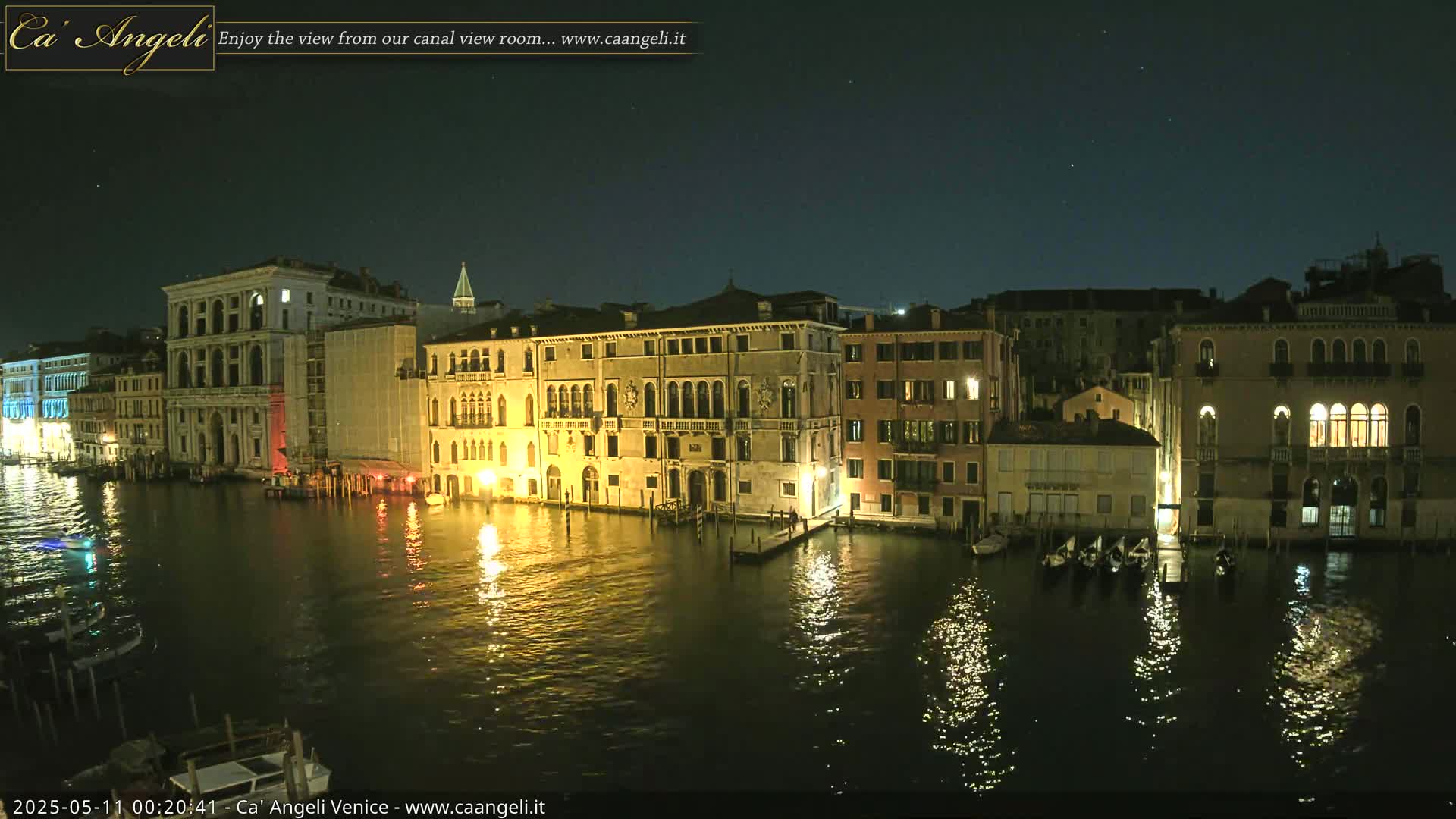 A nighttime view of illuminated buildings lining a canal, reflecting in the calm water under a clear, starry sky.