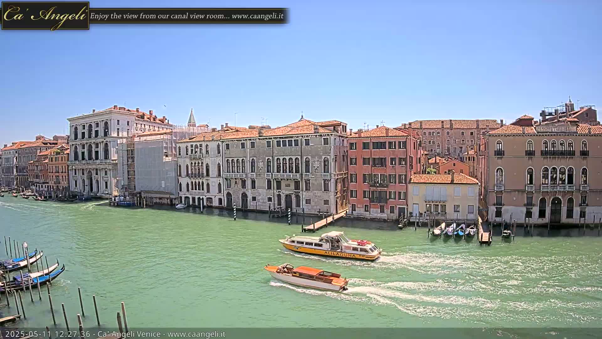 Under a clear blue sky, several water taxis and gondolas navigate a canal lined with historic Venetian buildings.