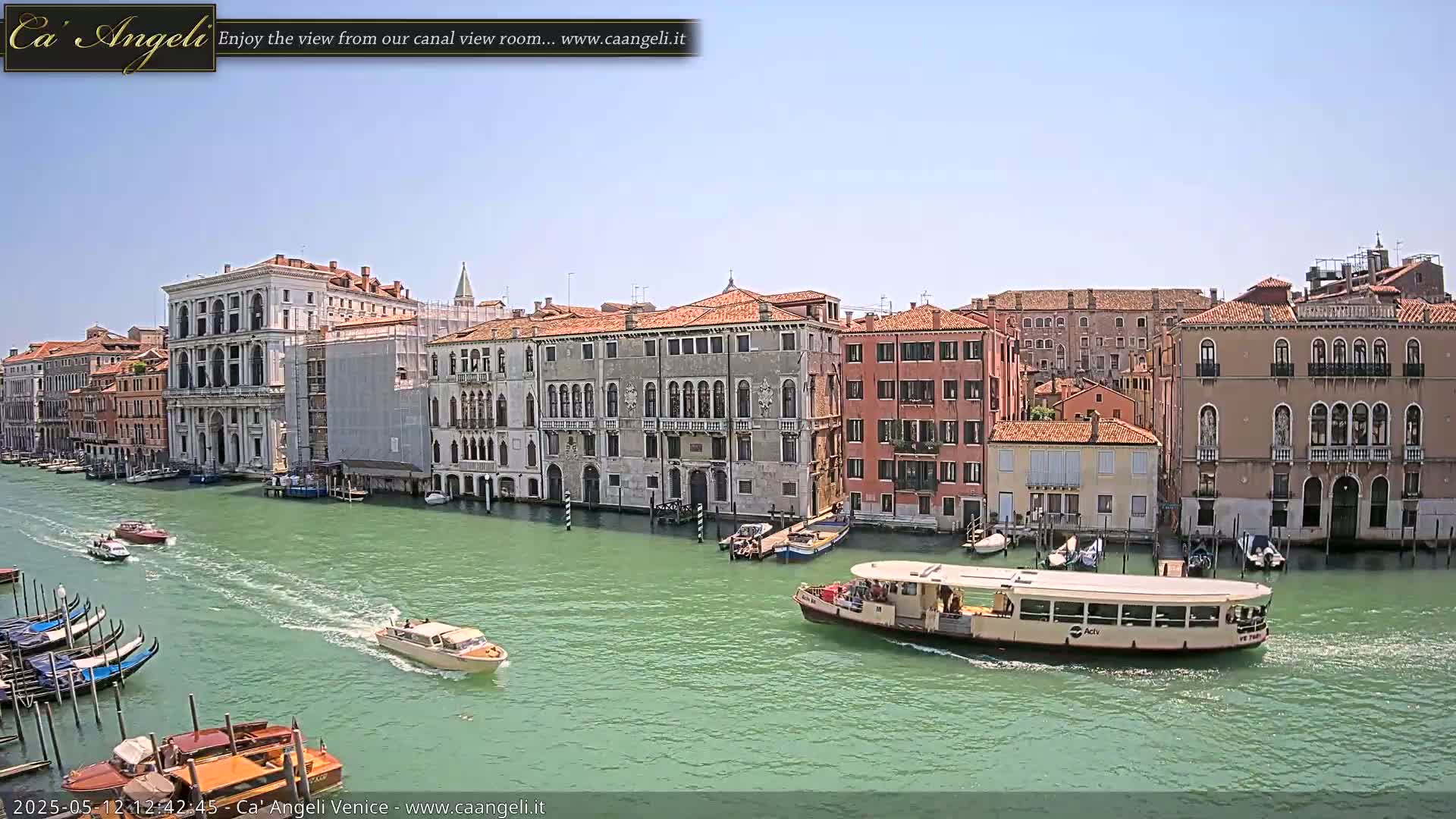A sunny day in Venice shows a canal with several boats, gondolas, and buildings lining the waterway.