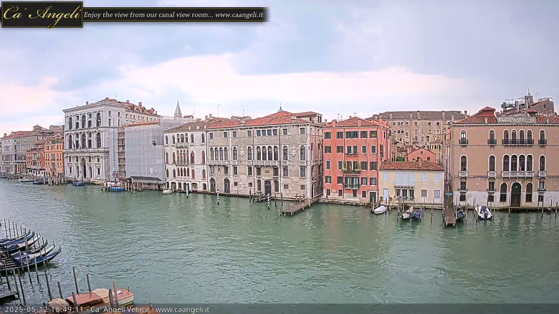 A wide shot shows a canal in Venice, Italy, under an overcast sky, with numerous buildings lining the waterway and gondolas moored along the banks.
