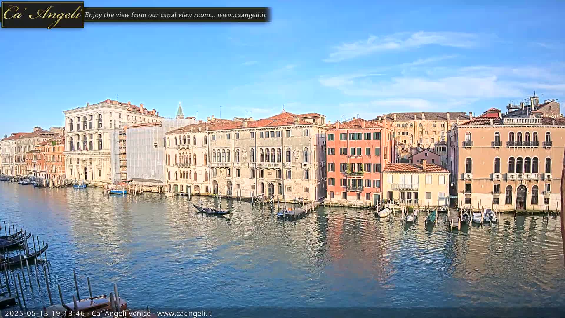 A sunny day in Venice shows a wide canal with gondolas and several buildings along its edge, some of which are undergoing renovations.