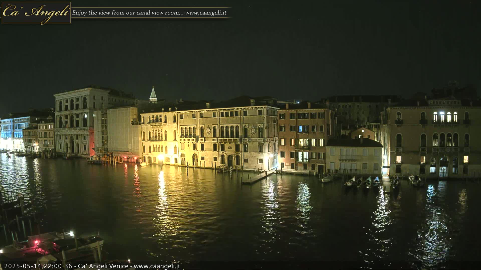 Nighttime view of illuminated buildings lining a canal, with several gondolas moored along the waterfront under a clear, dark sky.