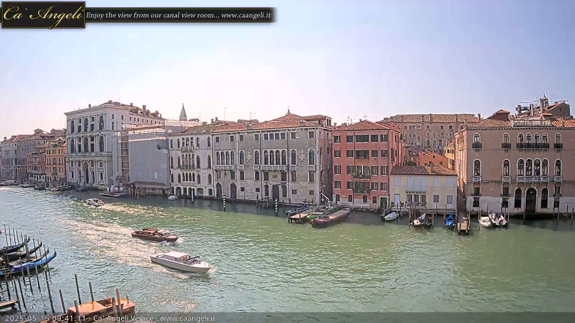 A sunny day in Venice shows several canal-side buildings, gondolas, and motorboats on a waterway.