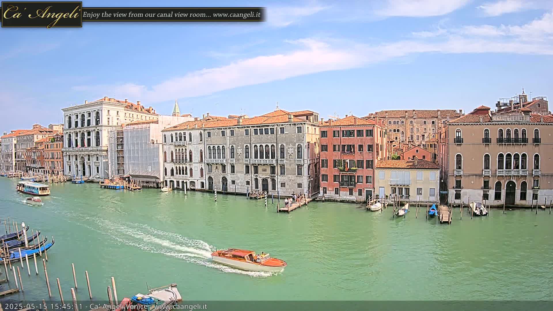 A canal in Venice, under a mostly sunny sky, is lined with historic buildings, and several boats are on the water.