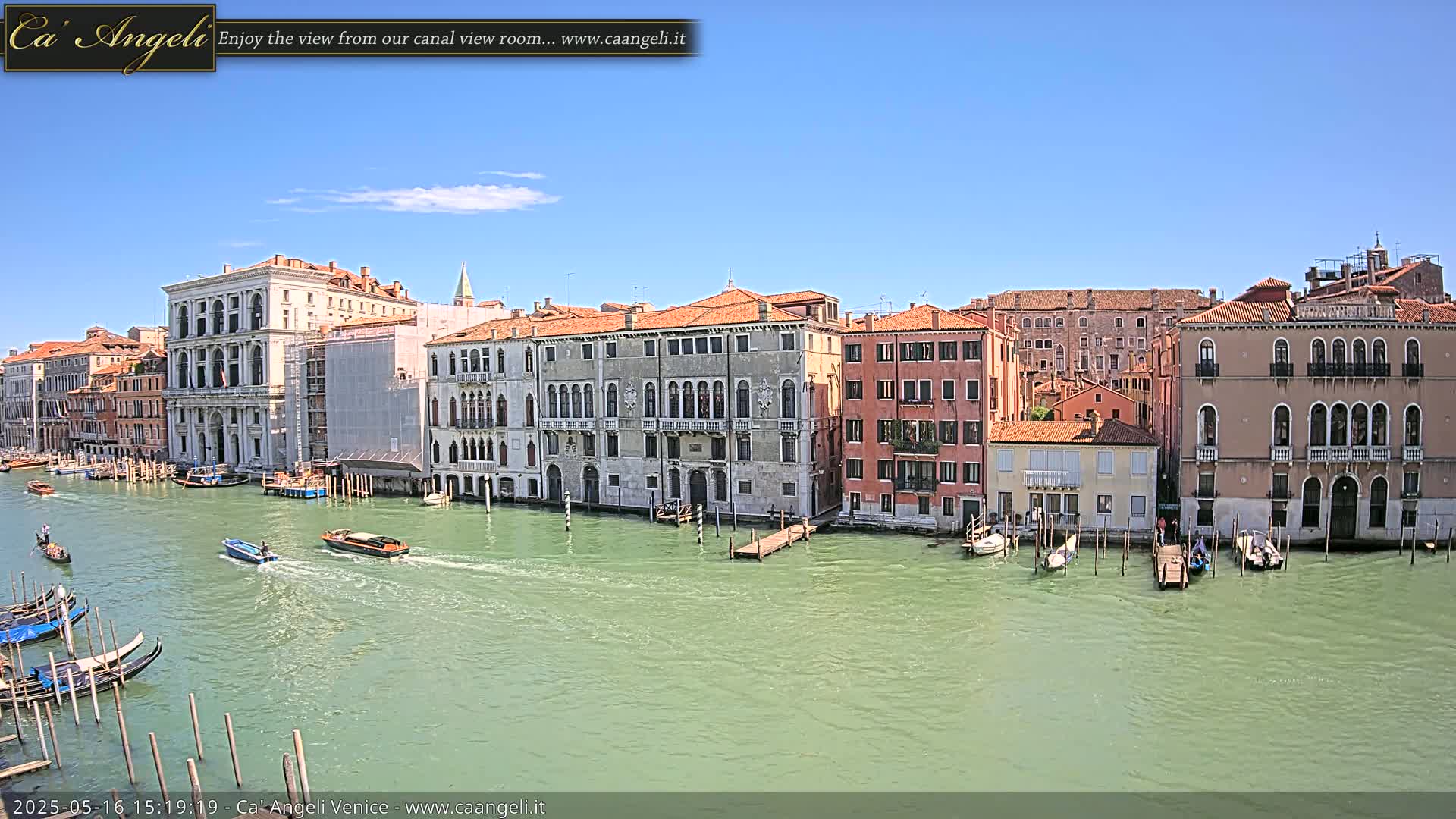 A sunny day in Venice shows a canal with gondolas and other boats passing by several historic buildings along the waterway.