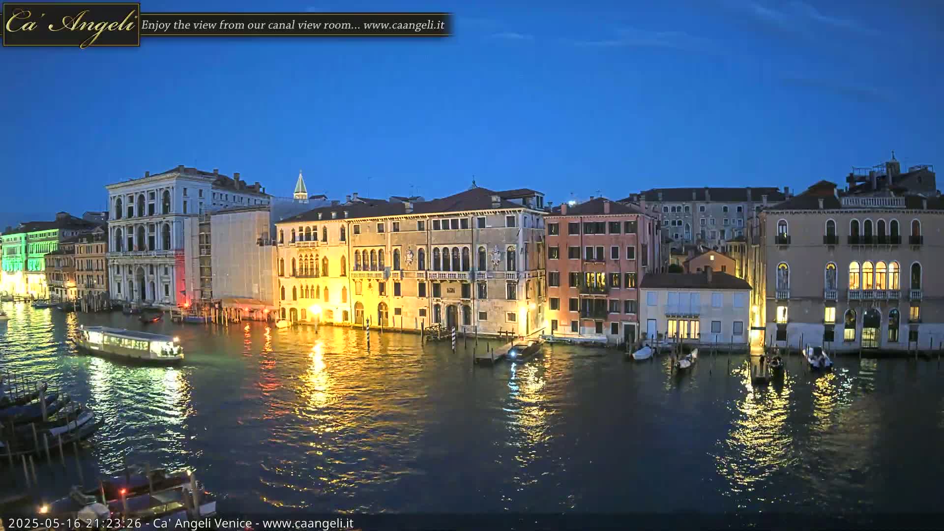 A tranquil canal scene at twilight in Venice, showing illuminated buildings lining the waterway with boats moored along the edges.