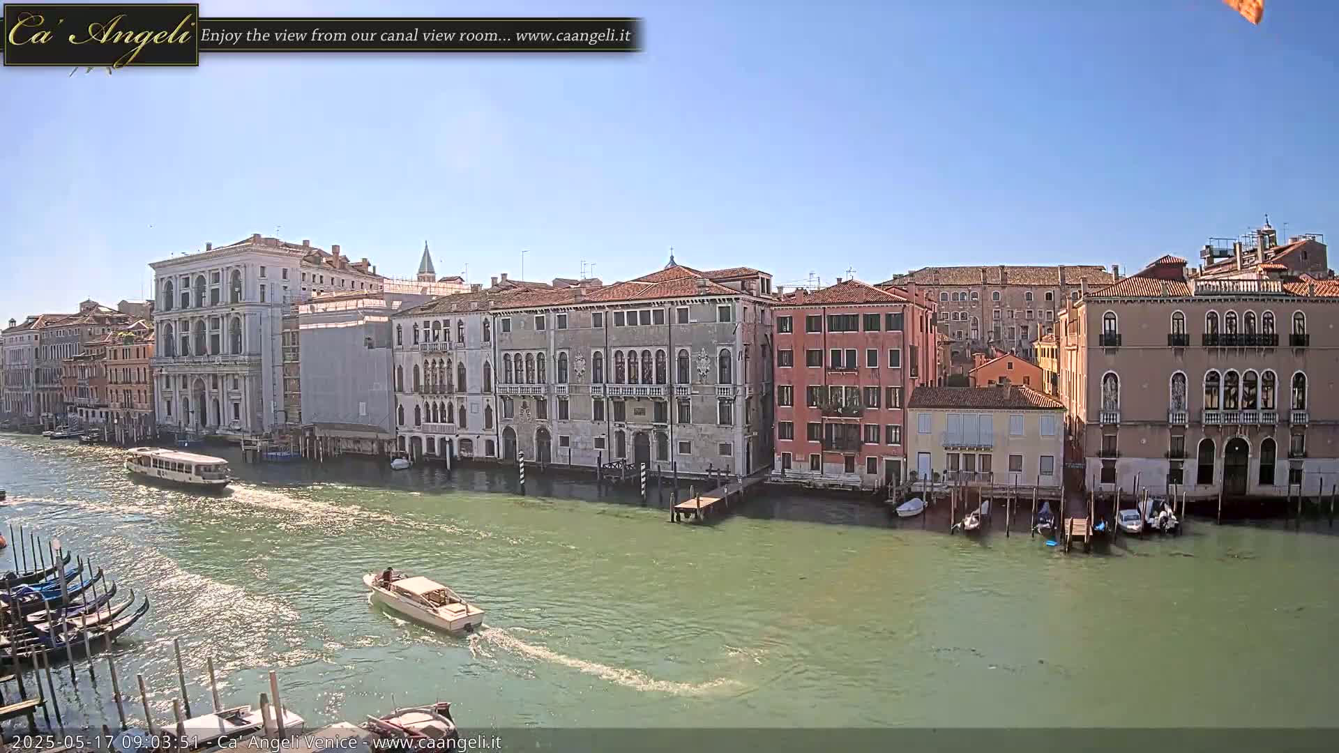 A sunny day in Venice shows a canal with several gondolas, a motorboat, and numerous buildings lining the waterway.