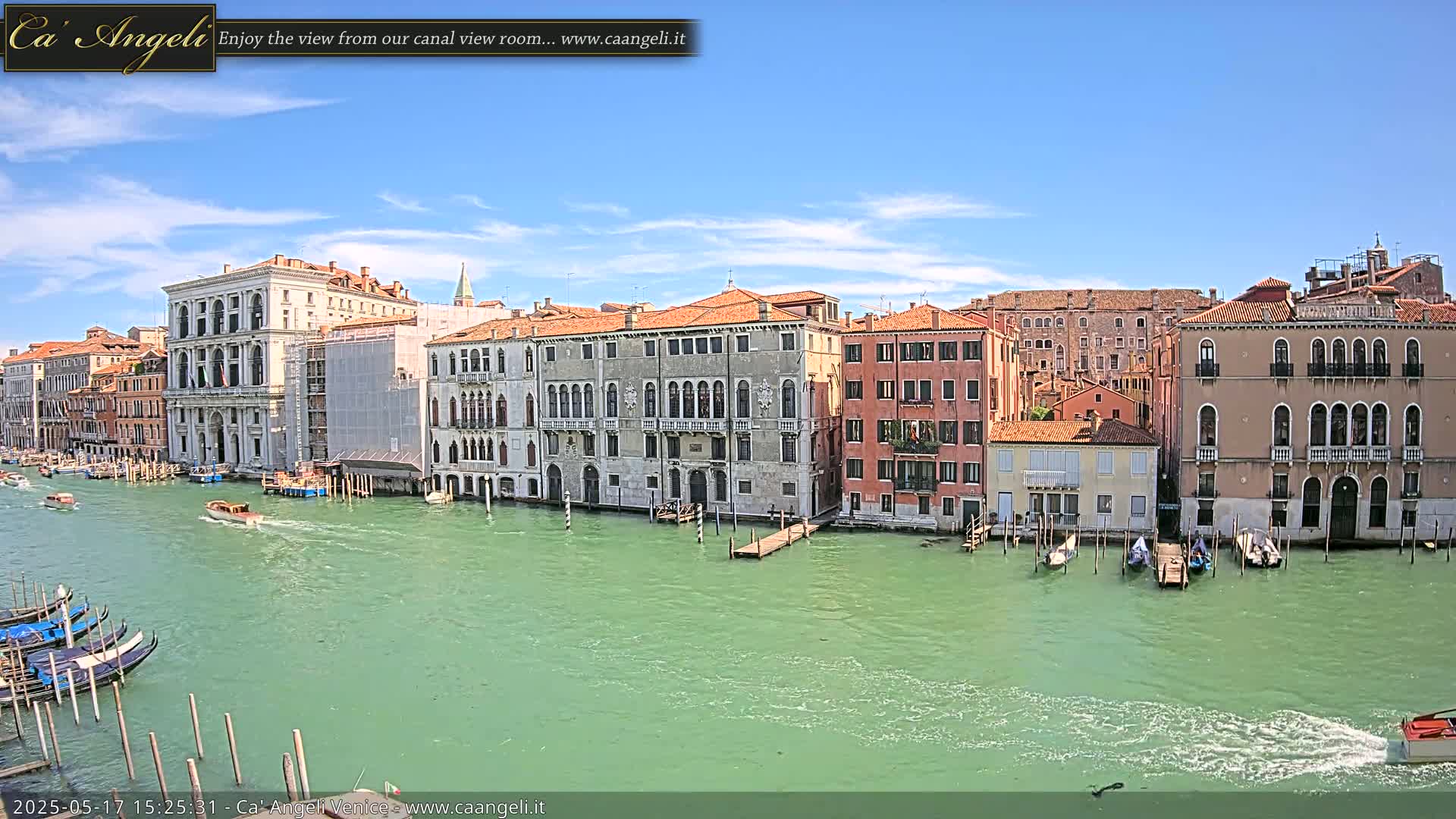 A wide canal in Venice, under a mostly sunny sky with a few clouds, is lined with several historic buildings, with boats and gondolas on the water.