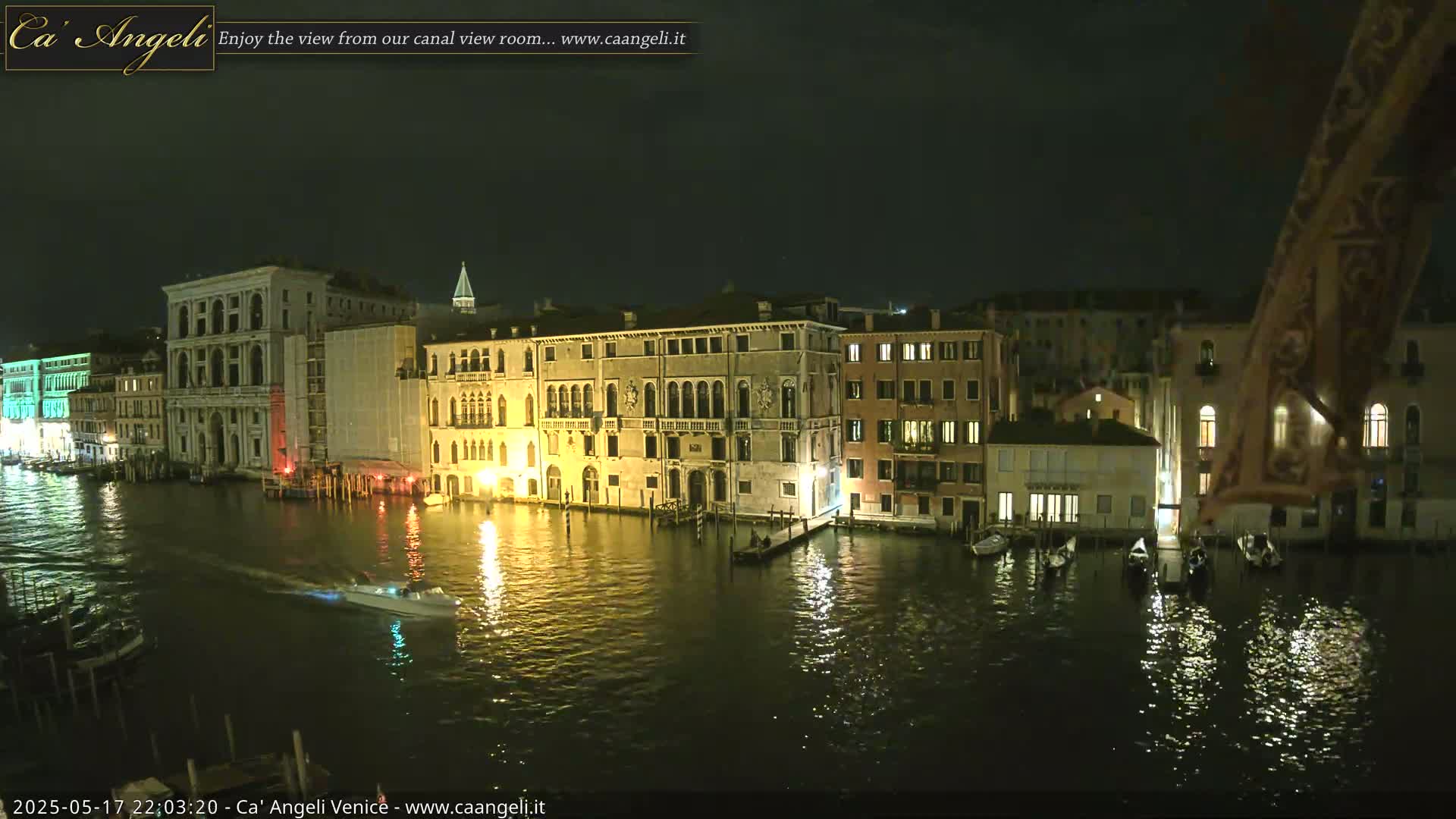 A nighttime view of a canal in Venice, Italy, shows several illuminated buildings lining the waterway, with a few boats moored along the edge and a small motorboat traversing the calm water.