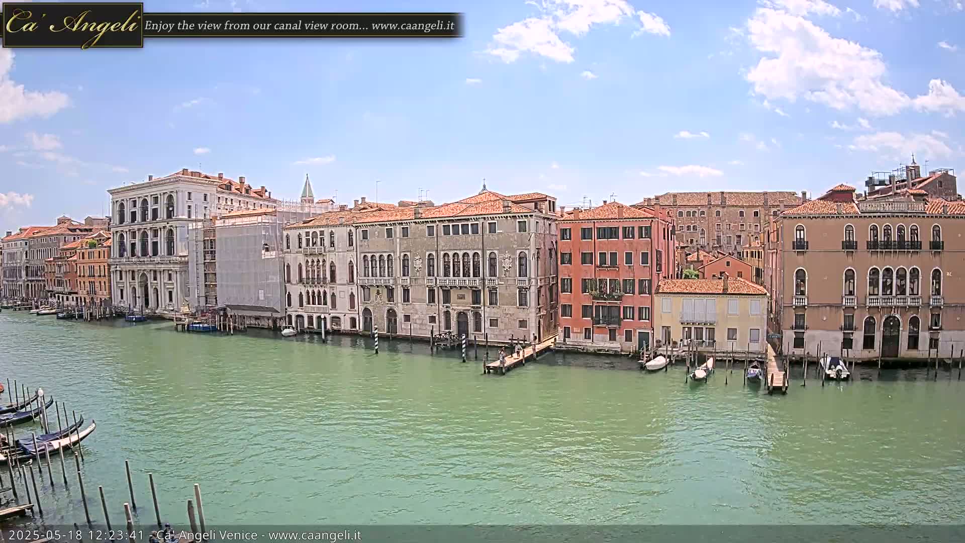 A canal in Venice, under a partly cloudy sky, is lined with several historic buildings, some undergoing renovations, with gondolas moored along the waterway.