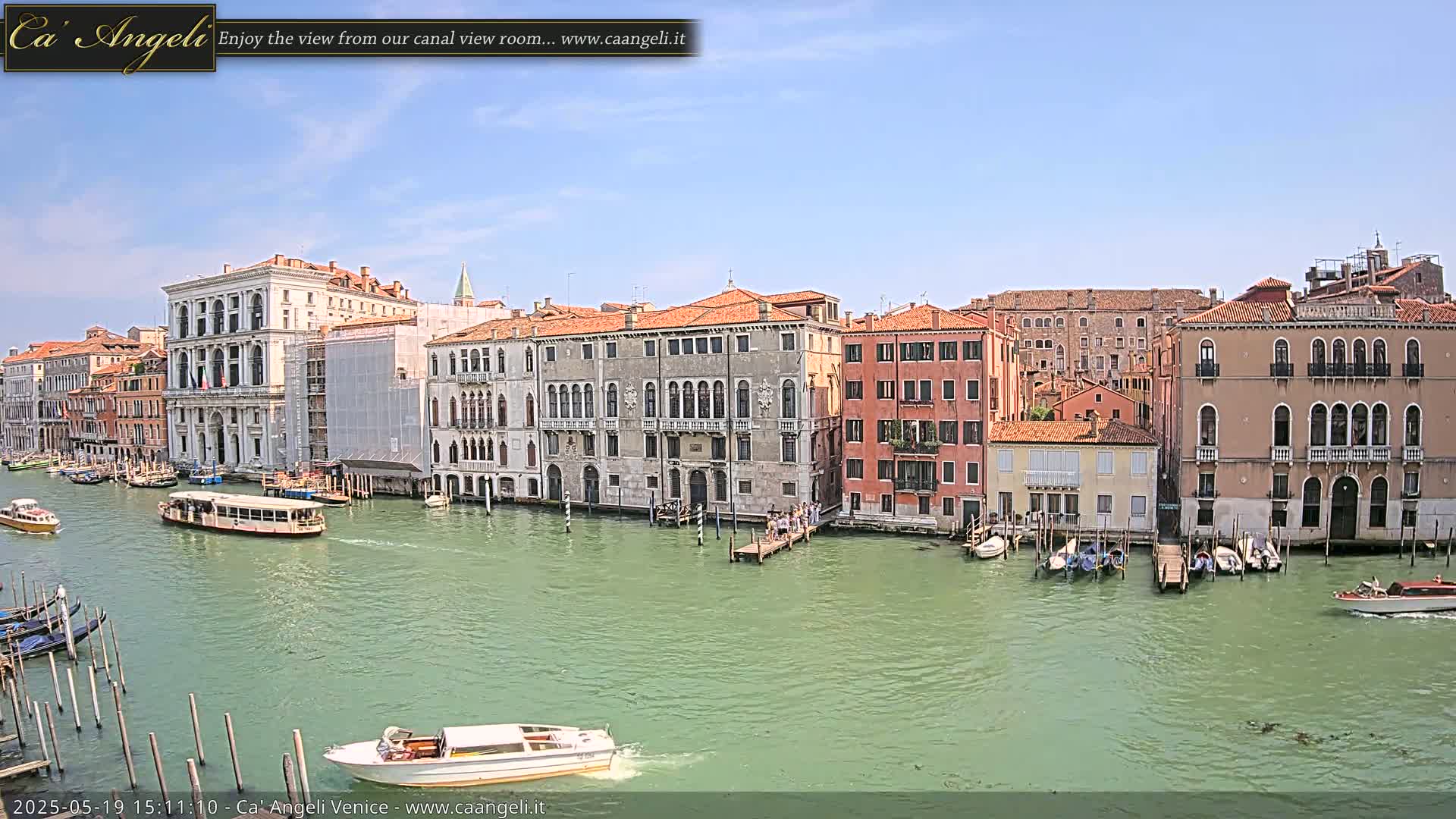 A sunny day in Venice shows a wide canal with several boats, gondolas, and buildings lining the waterway.