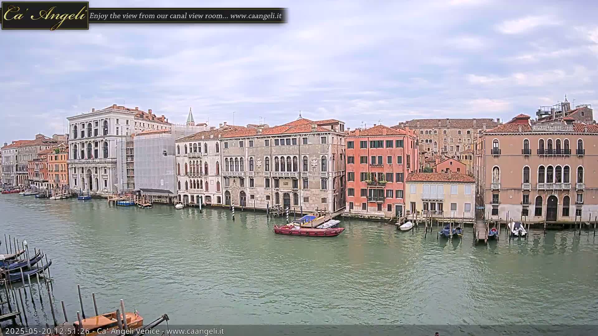 A waterway lined with Venetian buildings under a partly cloudy sky shows several boats moored along the canal's edge.