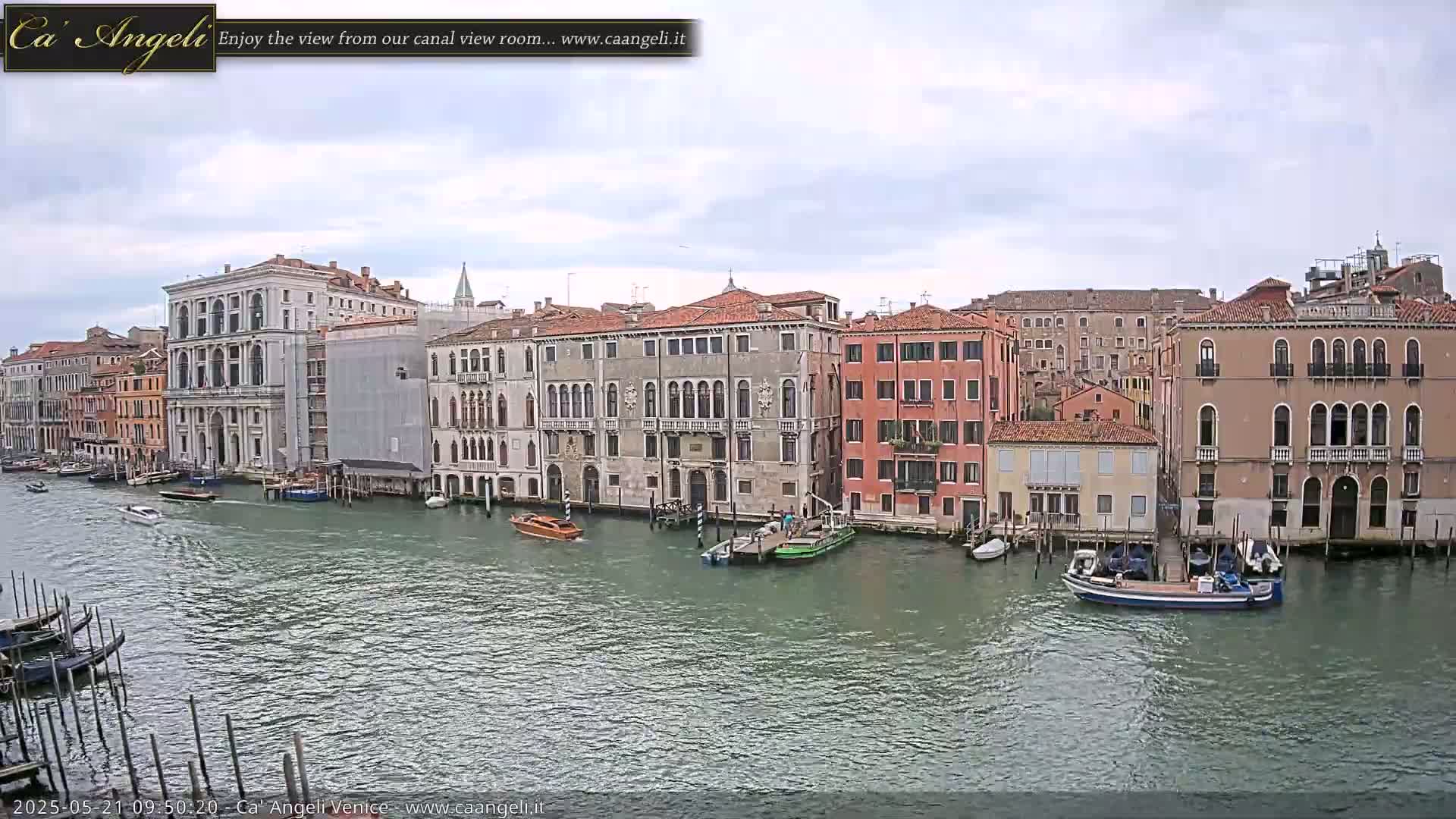 A wide shot of a canal in Venice, Italy on an overcast day, shows several buildings lining the waterway with boats moored along the edge and some in the canal itself.