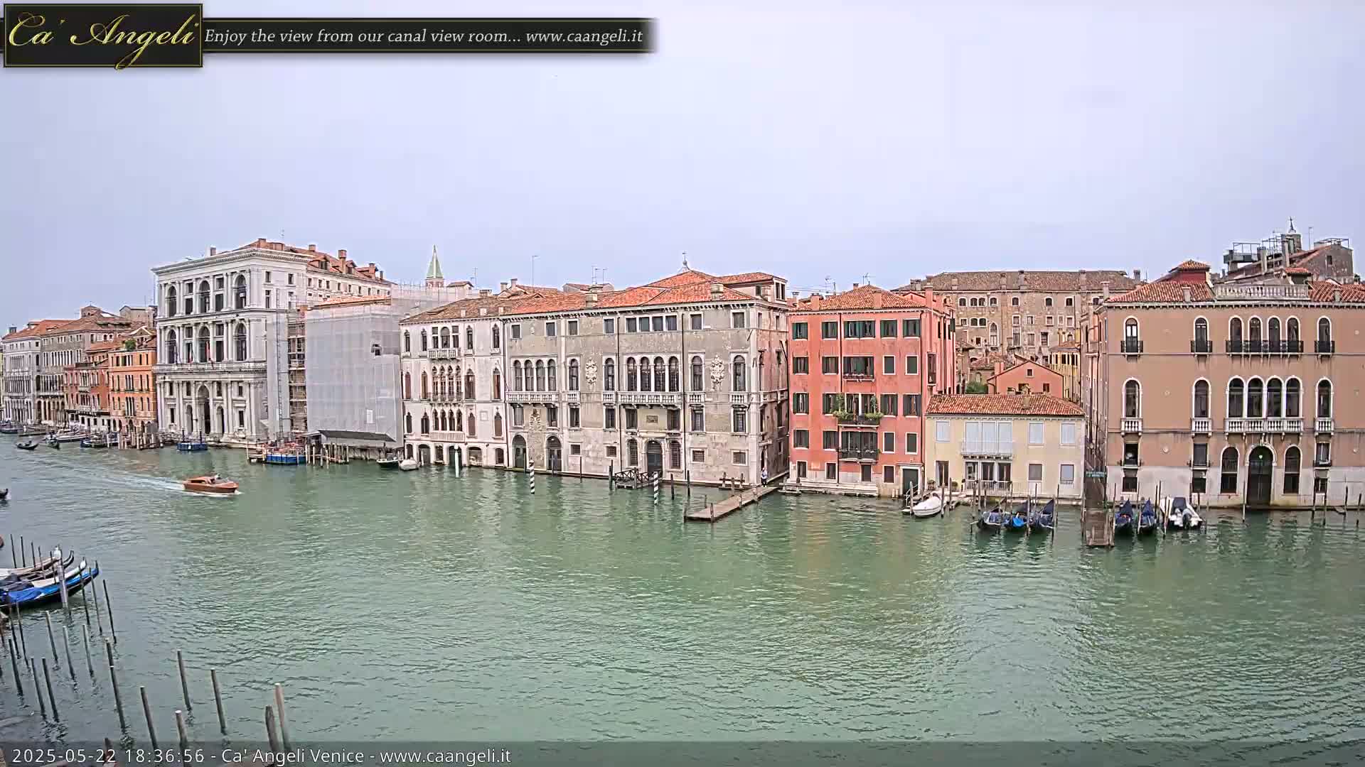 A canal in Venice, under an overcast sky, is lined with several multi-story buildings, some of which are under construction, with gondolas and motorboats on the water.